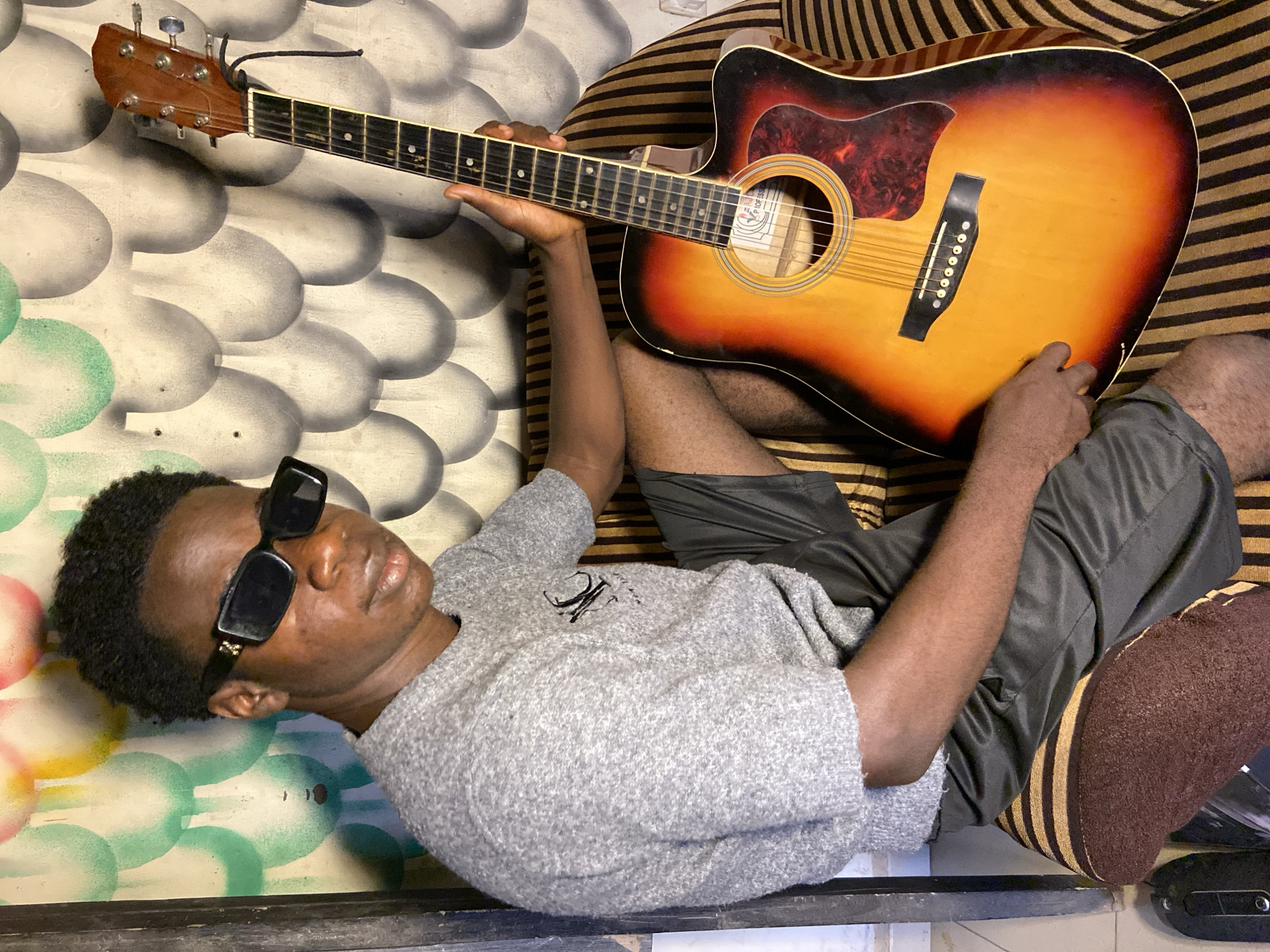An overhead shot shows a young man of dark complexion lying on his back, holding an acoustic guitar, in what appears to be an indoor setting. He has short, curly dark hair and is wearing black rectangular sunglasses, a light grey textured long-sleeved shirt, and dark shorts. His expression is relaxed, with his eyes seemingly closed or looking upwards behind the sunglasses.
The acoustic guitar features a sunburst finish, transitioning from dark brown edges to a bright orange-yellow center. It has a black pickguard and a dark fretboard with white dot inlays. His left hand is positioned on the guitar's neck, while his right hand rests near the bridge, suggesting he is either playing or posing with the instrument. Inside the soundhole, a label is partially visible, with the word "PROFESSIONAL" discernible.
The man is positioned against two different backdrops. To his left, there is a wall or surface with a repeating pattern of white raised oval shapes, partially painted in sections of light green and yellow. To his right and beneath the guitar, he is lying on a surface with alternating brown and yellow stripes, resembling a cushion or upholstered furniture. The overall mood of the image is relaxed and casual. There are no specific visual cues to definitively confirm the location as Mararaba, Nigeria, other than the provided context.