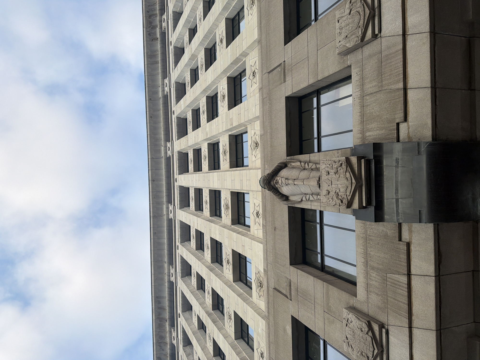 A low-angle shot looking up at a building with a repetitive window pattern on the facade. The building is made of gray stone or concrete. The windows are rectangular and dark, reflecting the sky. There are decorative carvings on the building’s exterior, including reliefs of figures and symbols. The sky is visible in the upper left corner, with scattered clouds.