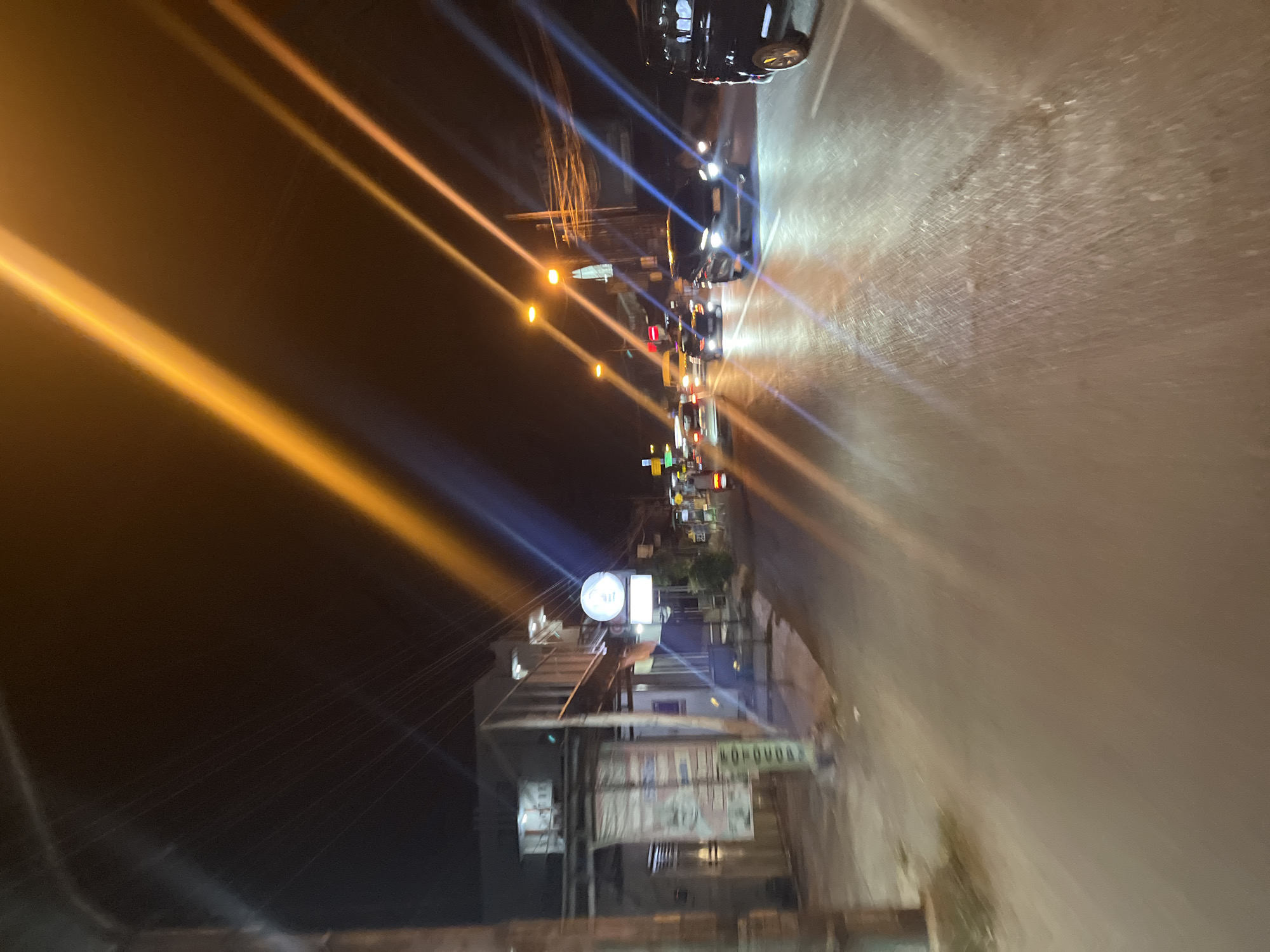 The image depicts a street scene at night in Kalinyamatan, Indonesia. The street is wet and reflects the headlights of passing vehicles, creating streaks of light across the pavement. Several cars are visible on the road, some with their headlights on. Buildings line the left side of the street, with some illuminated windows suggesting occupancy. A signpost is visible on the left, and what appears to be a large banner or advertisement is attached to a structure. Streetlights and power lines are also present, contributing to the overall nocturnal ambiance. The image is characterized by motion blur, indicating that it was taken from a moving vehicle. The foreground is dominated by the wet road surface, while the background shows the illuminated street and buildings.