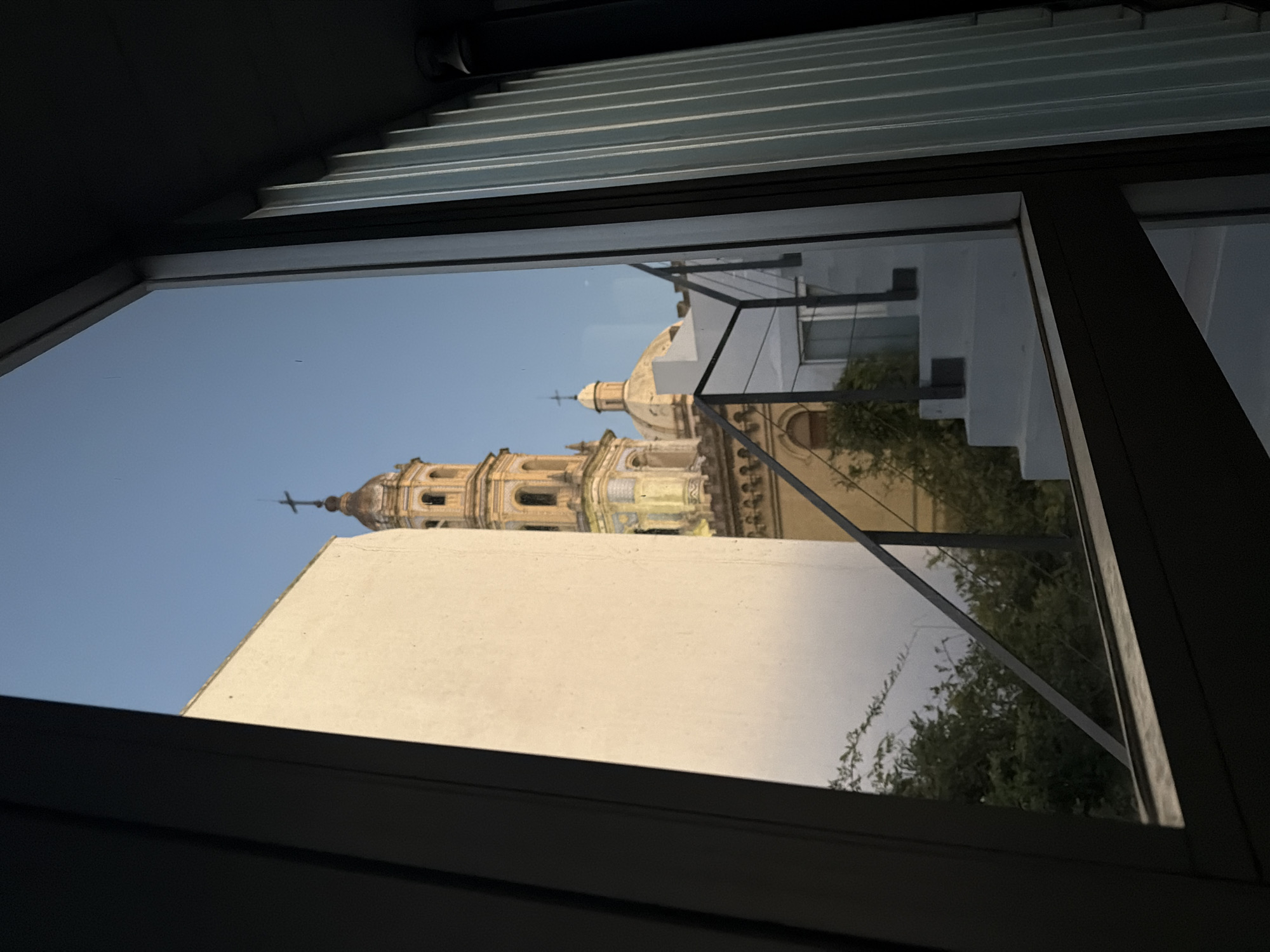 View of the tower of the Basilica of Our Lady of the Pillar from a window in Buenos Aires, Argentina. A white wall is visible in the foreground, obscuring the lower portion of the church. Trees are visible to the right of the frame. The sky is clear and light blue. The image is taken from an interior looking out, with a dark window frame and metal slats visible.