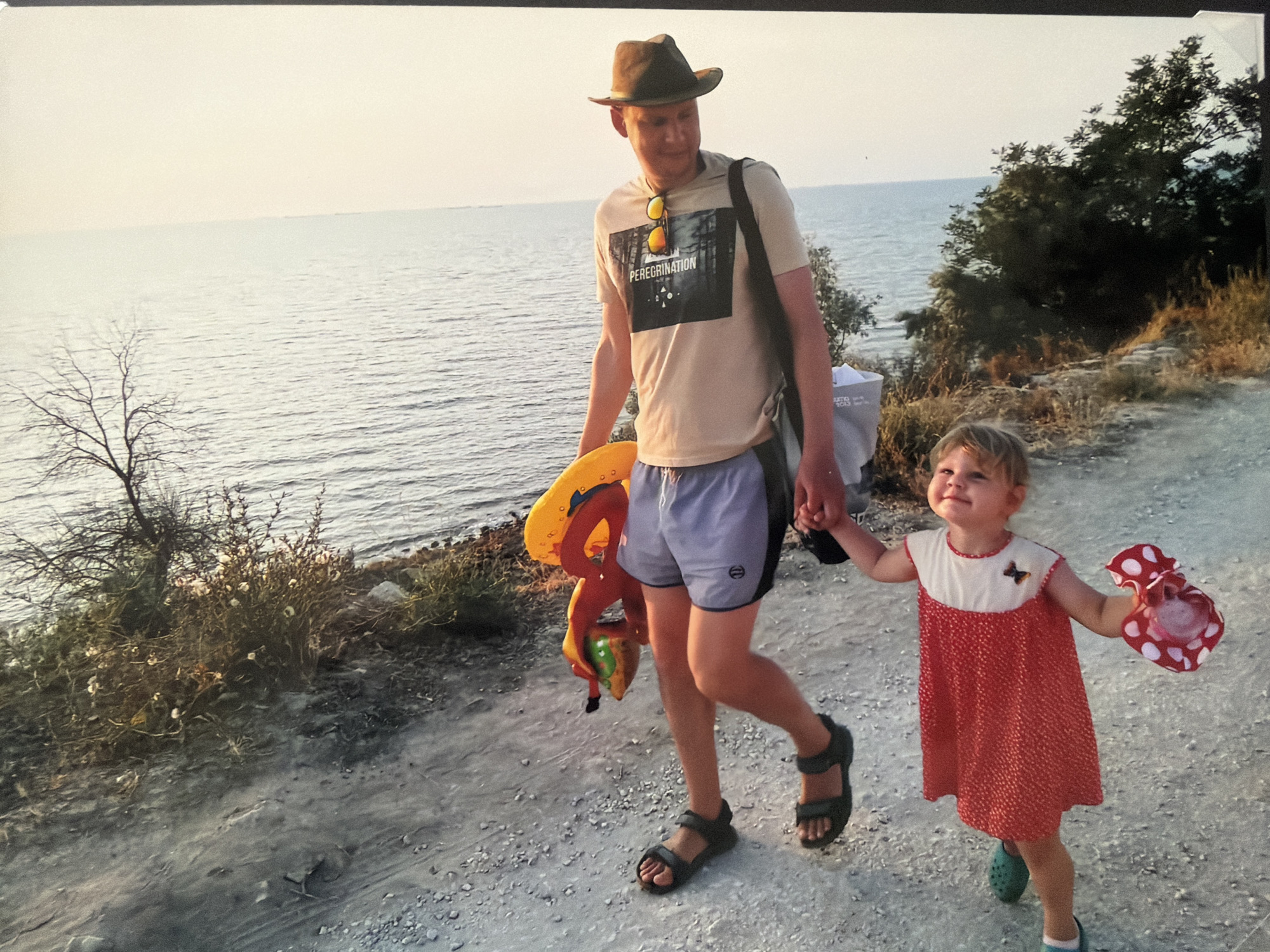 A medium shot depicts a man and a young girl walking hand-in-hand along a path near a body of water. The man, wearing a hat, T-shirt, shorts, and sandals, carries a yellow inflatable ring and other colorful inflatables in his left hand. Sunglasses are positioned on his shirt. He has a bag slung over his shoulder. To his right, a young girl in a red and white polka dot dress and Crocs holds what appears to be a red polka dot container in her free hand. The path they are walking on is made of small pebbles. To the left, sparse vegetation and a bare tree branch are visible. A dense thicket of trees and bushes is to their right. The water stretches out to the horizon in the background. The location is Unknown, Unknown.