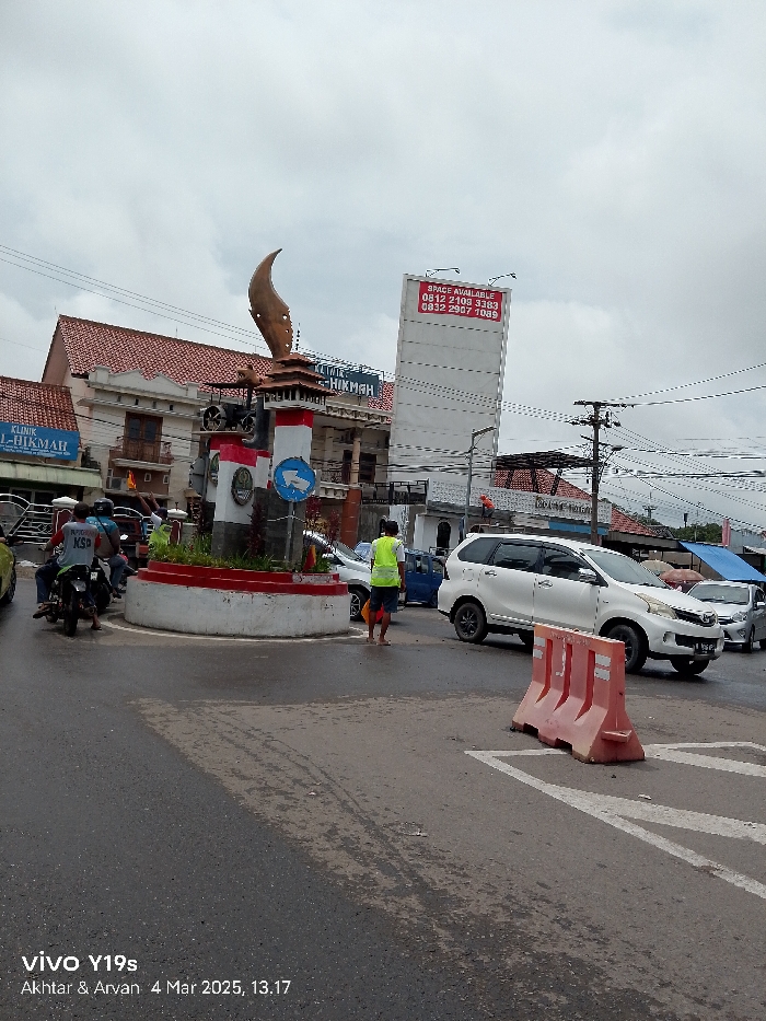 The scene is a street intersection in Jatibarang, Indonesia, taken on March 4, 2025, at 1:17 PM. A white, Toyota Avanza minivan is driving through the intersection. A man in a yellow vest is standing on the sidewalk, observing the traffic. The street is wet, indicating recent rain. A large, decorative monument is located in the center of the roundabout. Behind the monument is a two-story building with a sign that reads "Klinik Hikmah". There is a large white billboard to the right of the roundabout with the text “Space Available 0812 2109 3363 0832 2907 1089” on it. There is a small, blue car in the left lane. Two men on motorcycles are waiting at the intersection. There is a second, smaller billboard in the background, just visible above the white minivan. The traffic signal on the monument indicates a green light for traffic passing straight ahead. There is a large orange traffic barrier located in the right lane. The sky is grey and overcast, but there is no precipitation falling. The image was taken with a Vivo Y19s phone. Akhtar and Arvan are credited with the photograph. The image is in a horizontal format. The camera is positioned at street level.