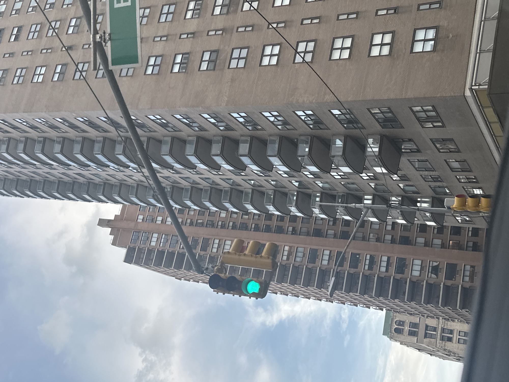 This is a medium shot of a city street in New York, United States of America. The main subjects are two tall apartment buildings, one brown brick and one beige brick. The brown brick building on the left is closer to the camera, and its facade is lined with balconies. The beige brick building on the right is further away, and its facade is also lined with balconies. There is a traffic light with a green light illuminated hanging from a wire, and a green street sign with white lettering that reads "E". The sky is cloudy and overcast. The image appears to have been taken from a vehicle.