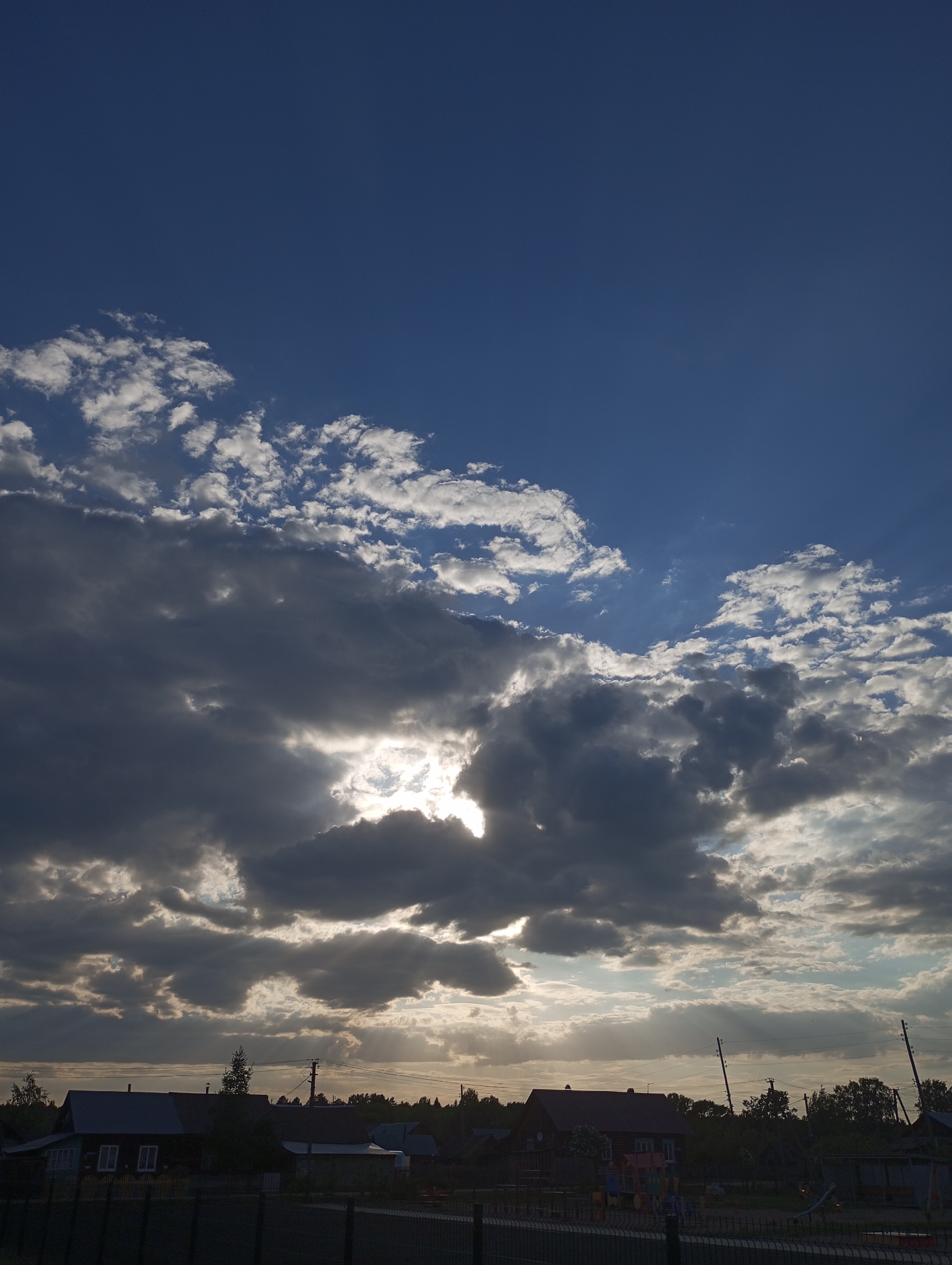 The image shows a cloudy sky over the city of Буренка, Russia. The sky is a mix of dark storm clouds and patches of blue sky. The sun is obscured by the clouds, but its rays are visible. In the foreground is a fence, beyond which are houses and trees.
