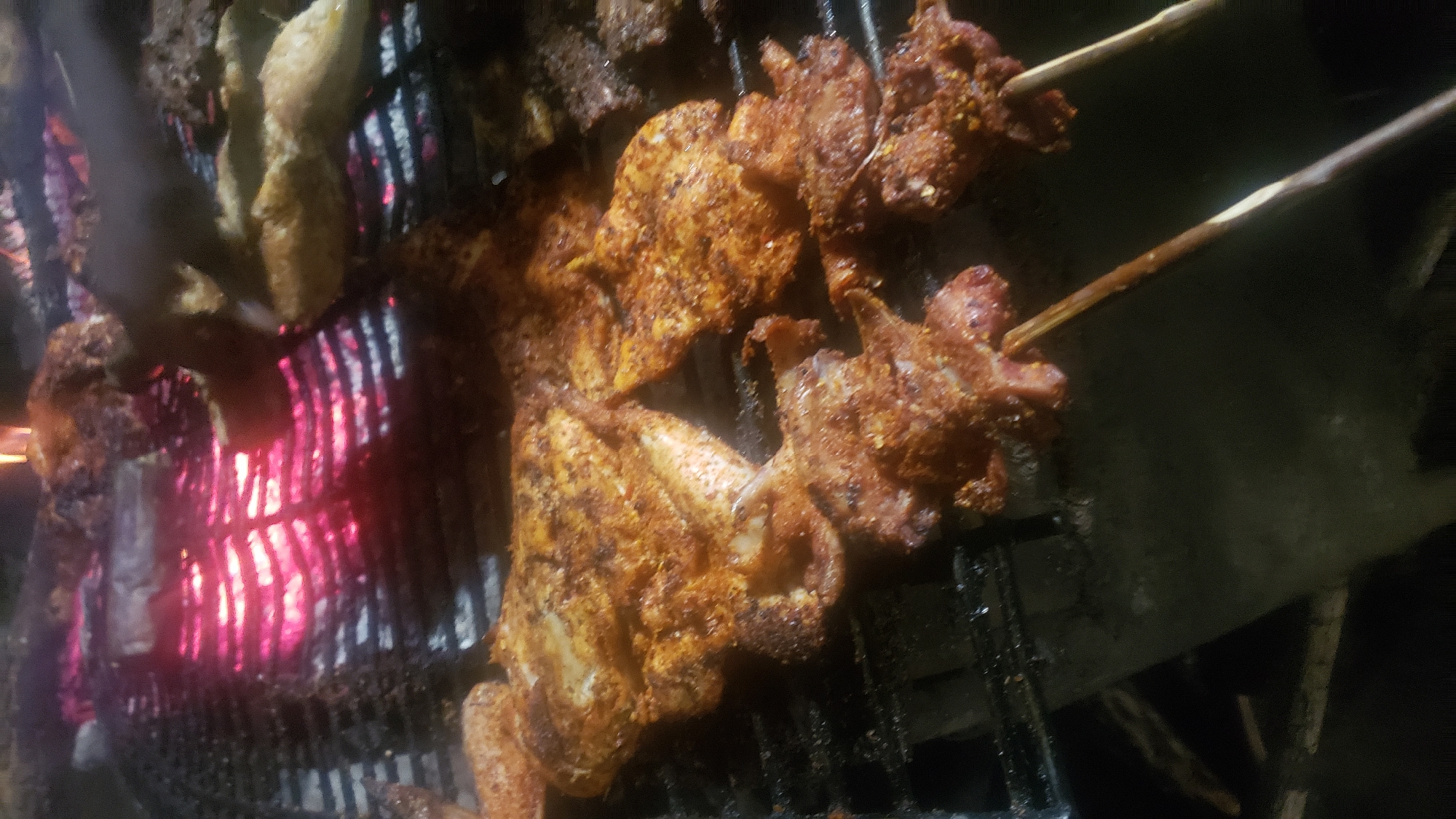 In Narayi, Nigeria, a close-up, low-angle shot captures grilled meat cooking over glowing red embers on a black metal grate. The meat appears to be seasoned with a reddish-brown spice blend. Some pieces are skewered on wooden sticks. The background is dark and out of focus, with hints of flames from the fire.
