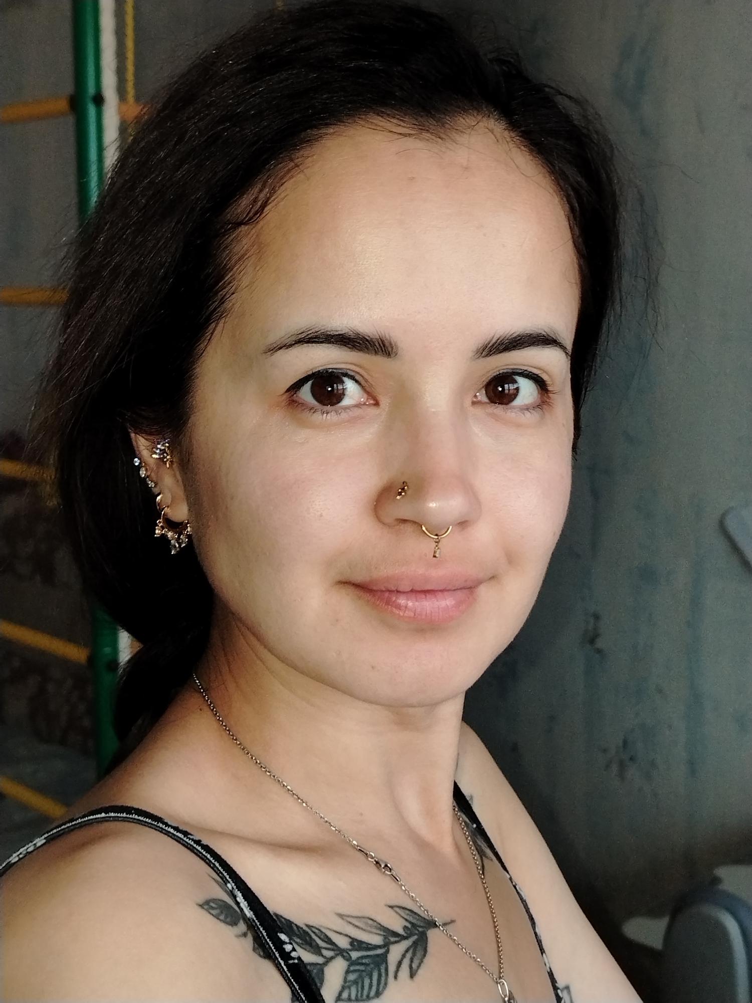 A headshot of a young woman with dark hair and brown eyes. She is wearing multiple earrings in her left ear and a septum ring. A necklace with a pendant is visible around her neck, and a tattoo of leaves and branches is visible on her chest. The background is a grey, textured wall with some vertical yellow and green bars to the left. The image does not provide any information about the city of Noviy, Russia.