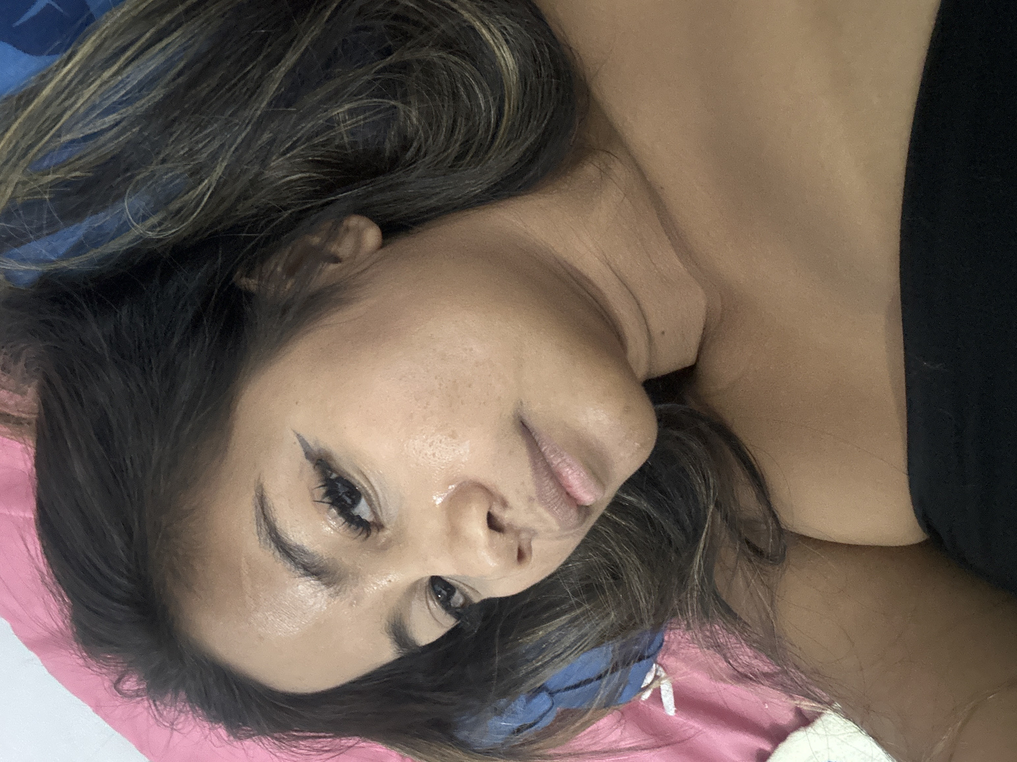 This is a close-up, sideways shot of a young woman lying on a pink surface, possibly a bed or cushion. She has long, dark brown hair with lighter highlights, which is spread around her head. Her face is turned towards the camera, with her left eye being more prominent. She appears to be wearing winged eyeliner and has well-defined eyebrows. There are some freckles visible on her cheeks and nose. Her lips are slightly parted, and she has a neutral expression. She is wearing a black top that reveals her shoulder and upper chest. The background shows hints of a blue fabric, possibly a pillow or blanket. The lighting is soft, suggesting an indoor setting, possibly during the day. There is no visible text in the image. The overall impression is of a casual, relaxed moment.