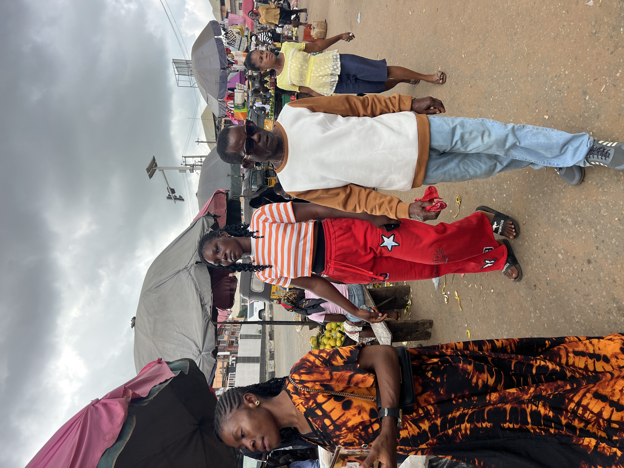 The image depicts a busy outdoor market scene in Nkpor, Nigeria, under an overcast sky. In the foreground, four people are walking. On the left, a woman with braided hair wears a vibrant orange and black patterned dress and carries a black purse. Next to her, a woman with braided hair walks in a bright orange and white striped t-shirt and red pants adorned with a white star. To their right, a man with sunglasses wears a white and brown sweatshirt and light blue jeans. Behind him, a woman in a yellow, white polka-dotted blouse and blue shorts walks away from the camera. Several market stalls with umbrellas are visible in the background, along with various goods, people, and vehicles. The ground is a dusty, reddish-brown surface, typical of many Nigerian markets.