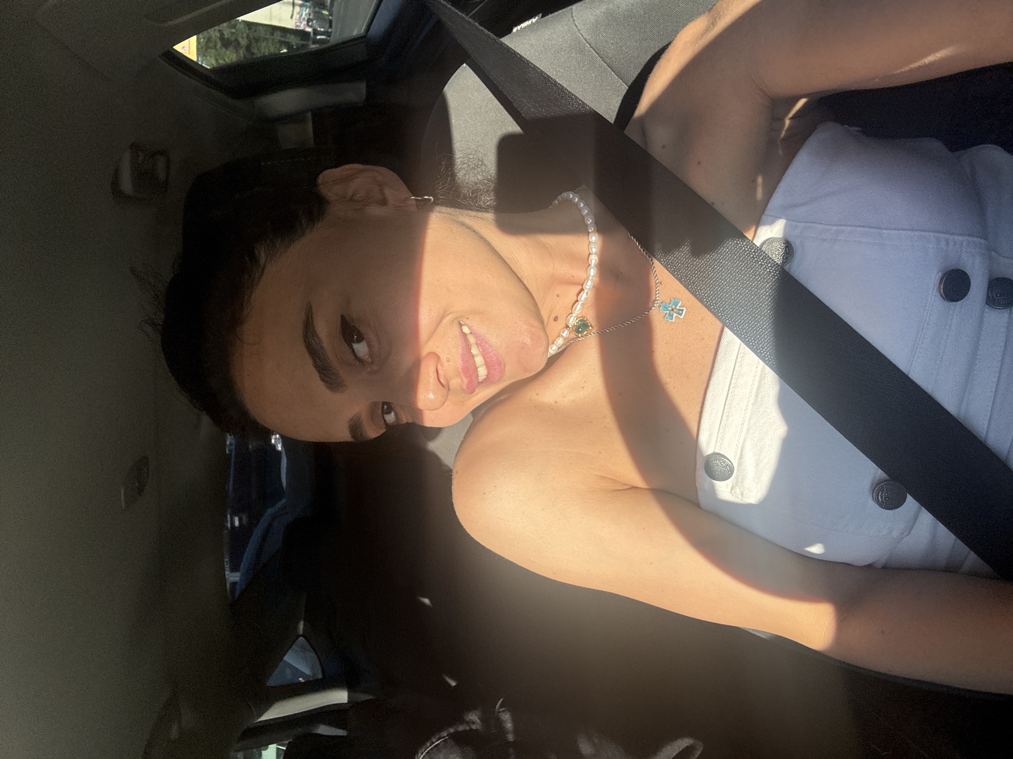 A woman is sitting in the passenger seat of a car, wearing a white strapless top and a pearl necklace with a green pendant and a blue cross pendant. She is smiling at the camera, and sunlight is streaming through the window, casting a shadow across her face and body. The interior of the car is visible, including the headrest and seatbelt. Outside the car window, trees and buildings can be seen, suggesting an urban environment. The location is identified as Tbilisi, Georgia.