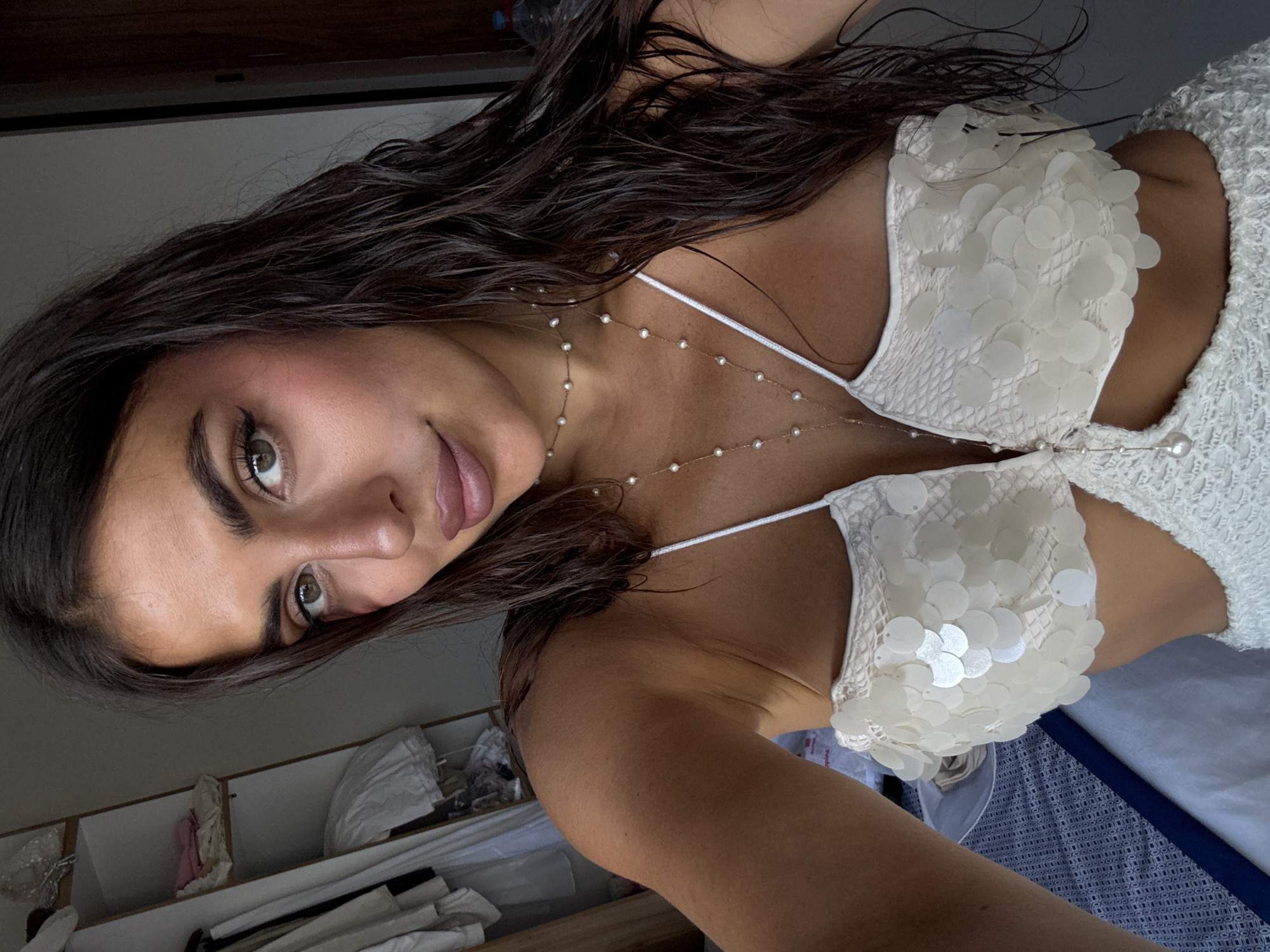 A woman with dark brown hair and light brown skin is indoors. She is wearing a white top with sequin details. She has a pearl necklace. Behind her is a white shelving unit with various items. The location of this image cannot be determined to be Kuta Selatan, Indonesia, without additional information.