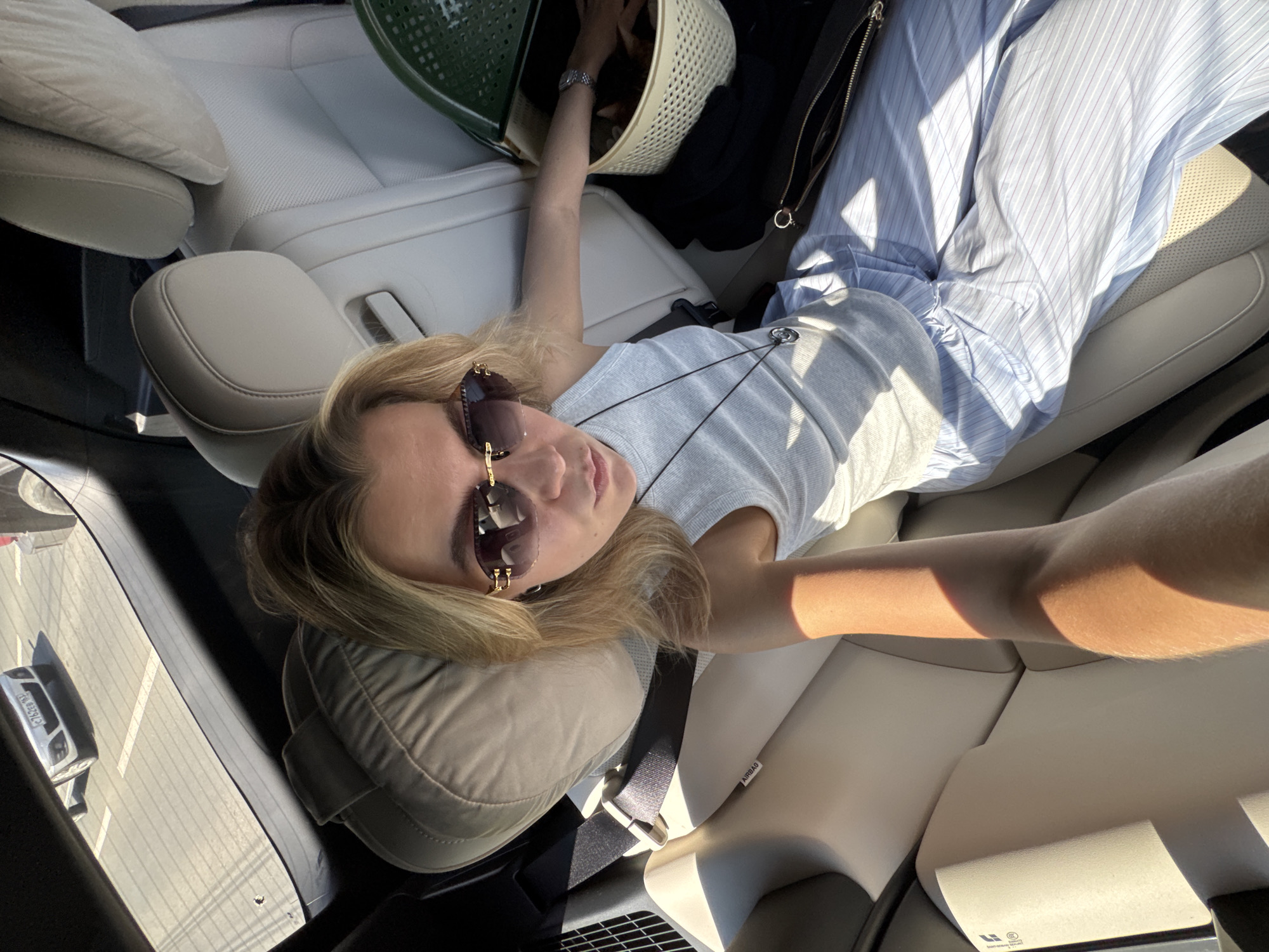 A woman with blonde hair, wearing sunglasses and a grey tank top, is sitting in the back seat of a car. She is wearing striped pants and a necklace. A basket filled with what appears to be fabric or yarn is next to her. Outside the car, a white car is parked in a parking lot. The scene is illuminated by bright sunlight.
