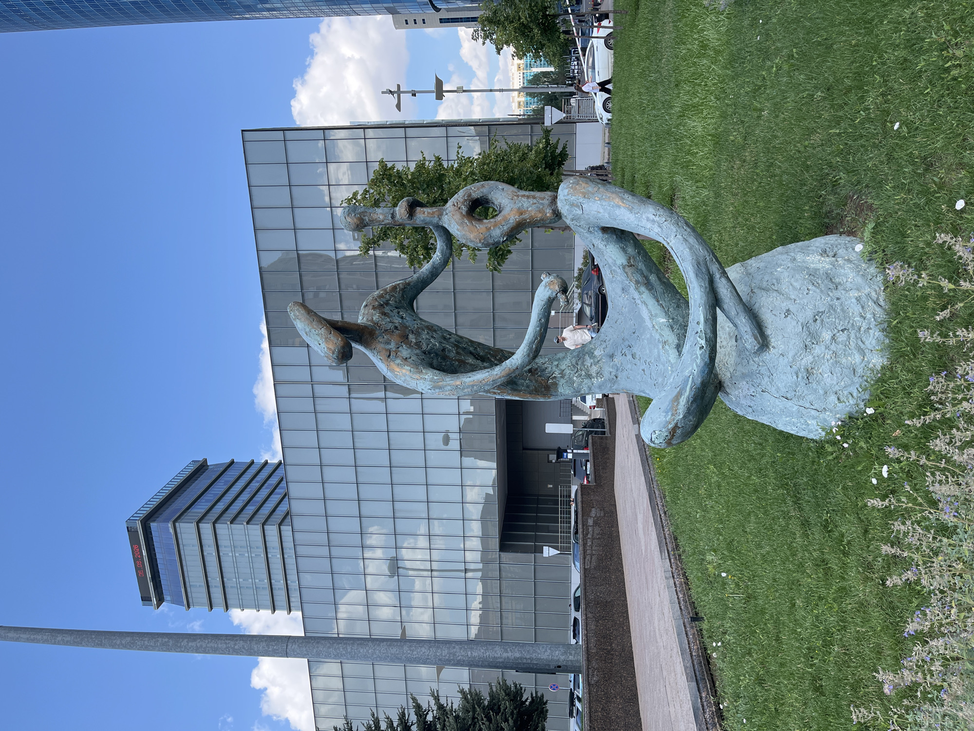 In Astana, Kazakhstan, a striking bronze sculpture depicting two abstract human-like figures in a seated, intertwined pose dominates the foreground of a grassy area. The sculpture rests on a rough, rock-like base. Behind the sculpture, a modern glass-fronted building with a grid-like facade reflects the sky and surrounding environment. To the left, a tall, gray pole extends vertically, obscuring part of the view of a multi-story building topped with a digital display showing "06.08.2026". Further to the right, another tall, blue-glass skyscraper pierces the sky. Several cars are visible parked and driving on a road in the mid-ground, adjacent to a paved walkway. Trees and streetlights are interspersed throughout the scene. The sky is a clear blue with scattered white clouds, indicating daytime.