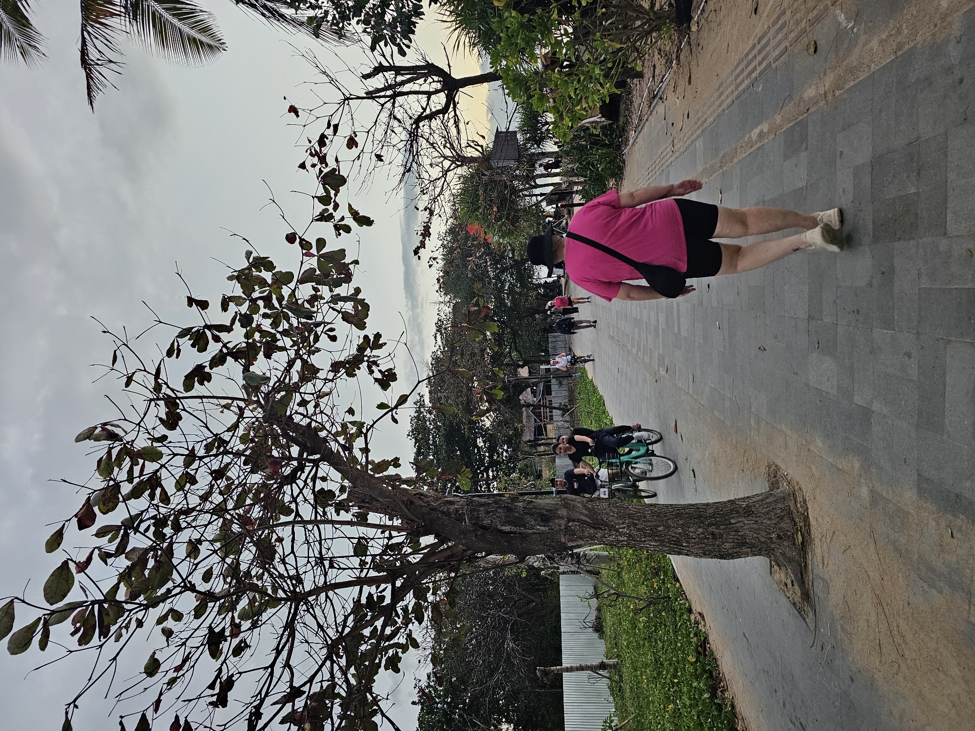 In Denpasar, Indonesia, a paved walkway runs alongside lush green vegetation and a metal fence. Several people are seen on the walkway: a woman in a pink t-shirt and black shorts walks away from the camera, carrying a black bag across her body; a couple on bicycles, a man and a woman, ride towards the foreground; and further down the path, other individuals can be seen cycling. The scene is framed by trees, some with sparse leaves and bare branches, and palm fronds are visible in the background. The sky is overcast with clouds.