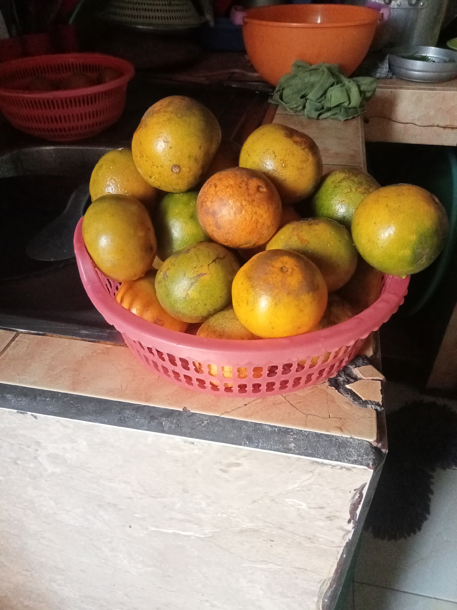 An overhead shot shows a pink plastic basket filled with oranges. The basket is on a counter that appears to be in a kitchen. There are other kitchen items in the background, including a red basket and an orange bowl. The scene is lit by natural light, suggesting it is daytime. The image is taken from a slight angle, with the basket of oranges being the main subject.