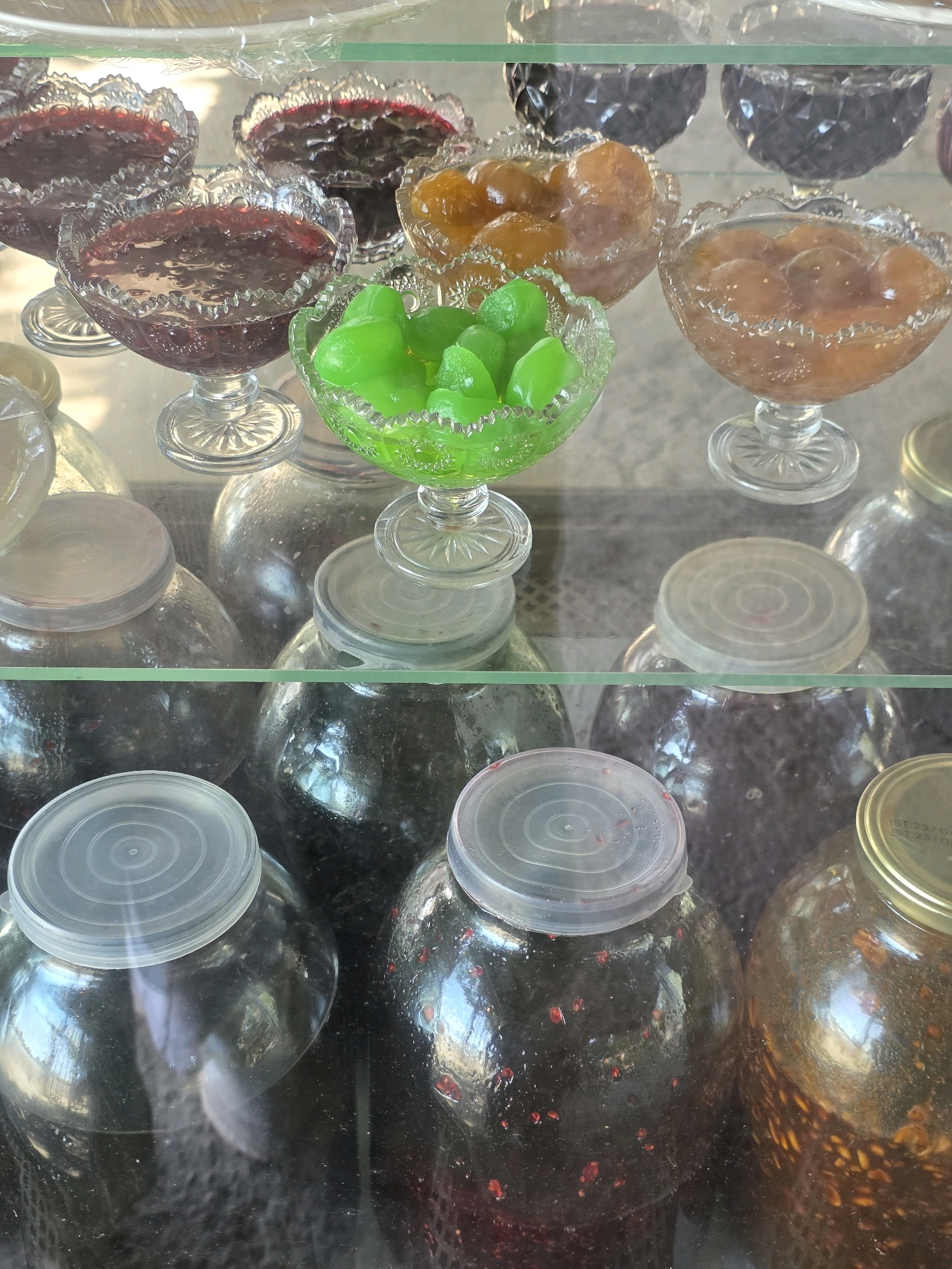 The image shows a display of preserved fruits and jams in glass containers, likely at a market stall or shop in Qobustan, Azerbaijan. In the foreground, several large glass jars with plastic or metal lids are filled with dark-colored preserved fruits or jams, some appearing to have whole berries or pieces of fruit. In the middle ground, smaller, decorative glass bowls hold preserved fruits. One bowl contains bright green, jelly-like pieces, while others hold preserved fruits in syrup, appearing amber or reddish-brown. In the background, more dark-colored preserved items are visible in decorative glass containers, some of which are partially obscured. The items are arranged on glass shelves, suggesting an indoor setting. The lighting is diffused, highlighting the textures and colors of the preserved items. There are no people visible in the image.