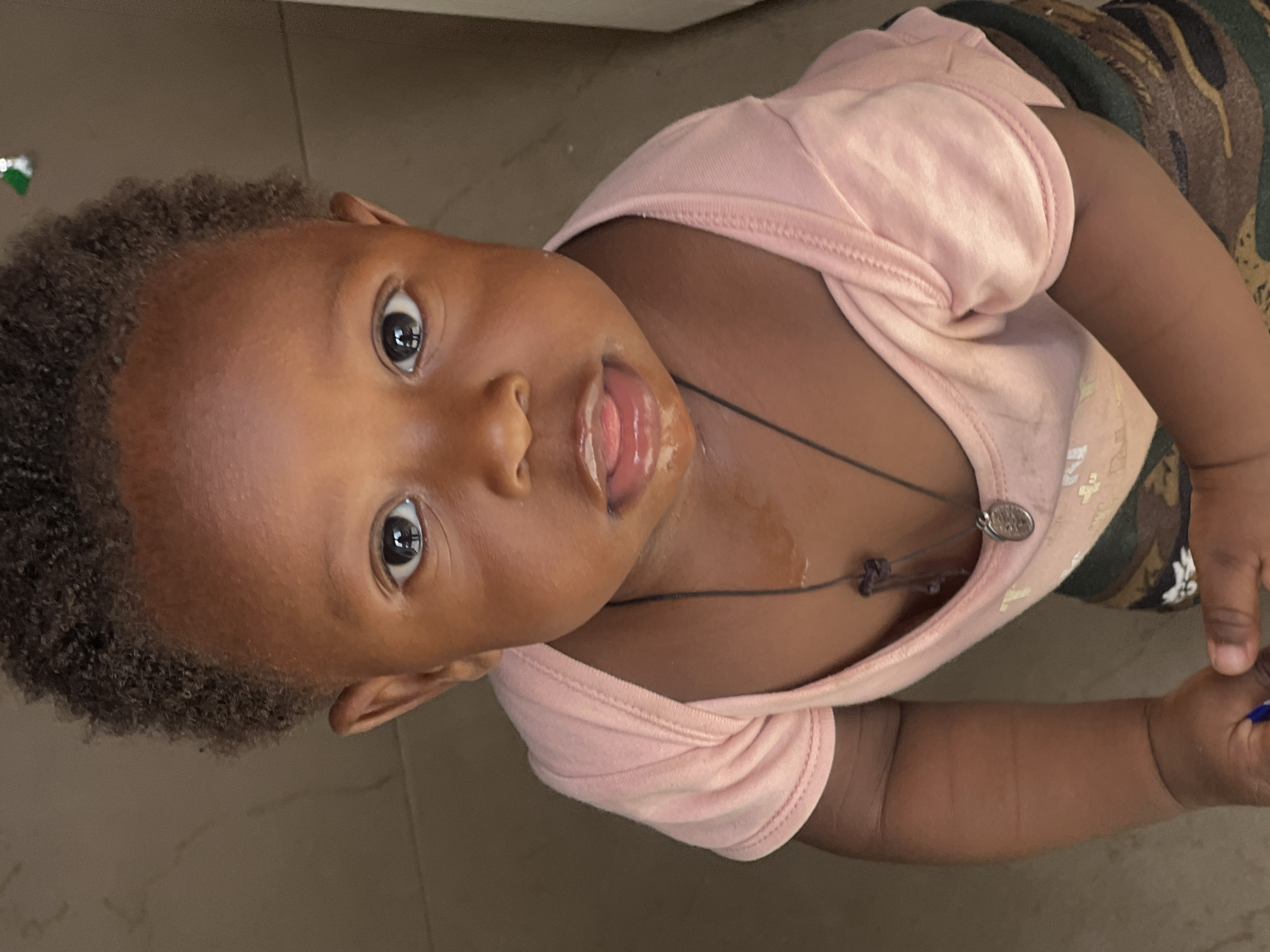A baby with brown skin, black curly hair, and large brown eyes is lying on a tan tile floor. The baby is wearing a light pink t-shirt and a camouflage print garment. A thin black necklace with a pendant is visible around the baby's neck. The baby's mouth is open and the tongue is visible. The baby's location is the city of Gura Top, Nigeria. A small blue object is being grasped by the baby in their left hand. A small fragment of a wrapper is visible in the upper left corner of the image.