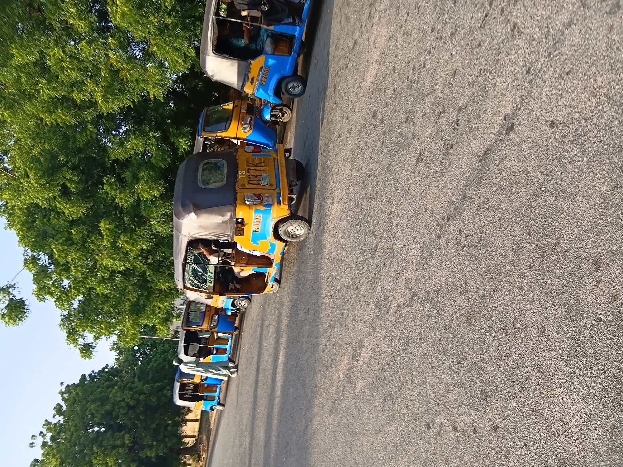 In Jalingo, Nigeria, a line of blue and yellow auto-rickshaws, known locally as "Keke napep" or "Maruwa," are parked on an asphalt road. The vehicles are positioned at an angle, with their fronts facing towards the left side of the frame. The road appears to be a well-maintained street, possibly in a residential or commercial area, with a sidewalk and trees lining the background. The sky above is clear and blue, indicating fair weather.
The auto-rickshaws are a common mode of public transportation in Nigeria. They are three-wheeled vehicles, powered by a small engine, and are designed to carry passengers. The rickshaws in the image are brightly colored, with blue bodies and yellow roofs. Some of them have people inside, suggesting they are either waiting for passengers or have just arrived at their destination.
In the foreground, a person wearing a light-colored traditional outfit is standing near the auto-rickshaws, possibly a driver or a passenger. The person is facing away from the camera, and their presence adds a human element to the scene. The auto-rickshaws are neatly parked, and the overall impression is one of order and activity.