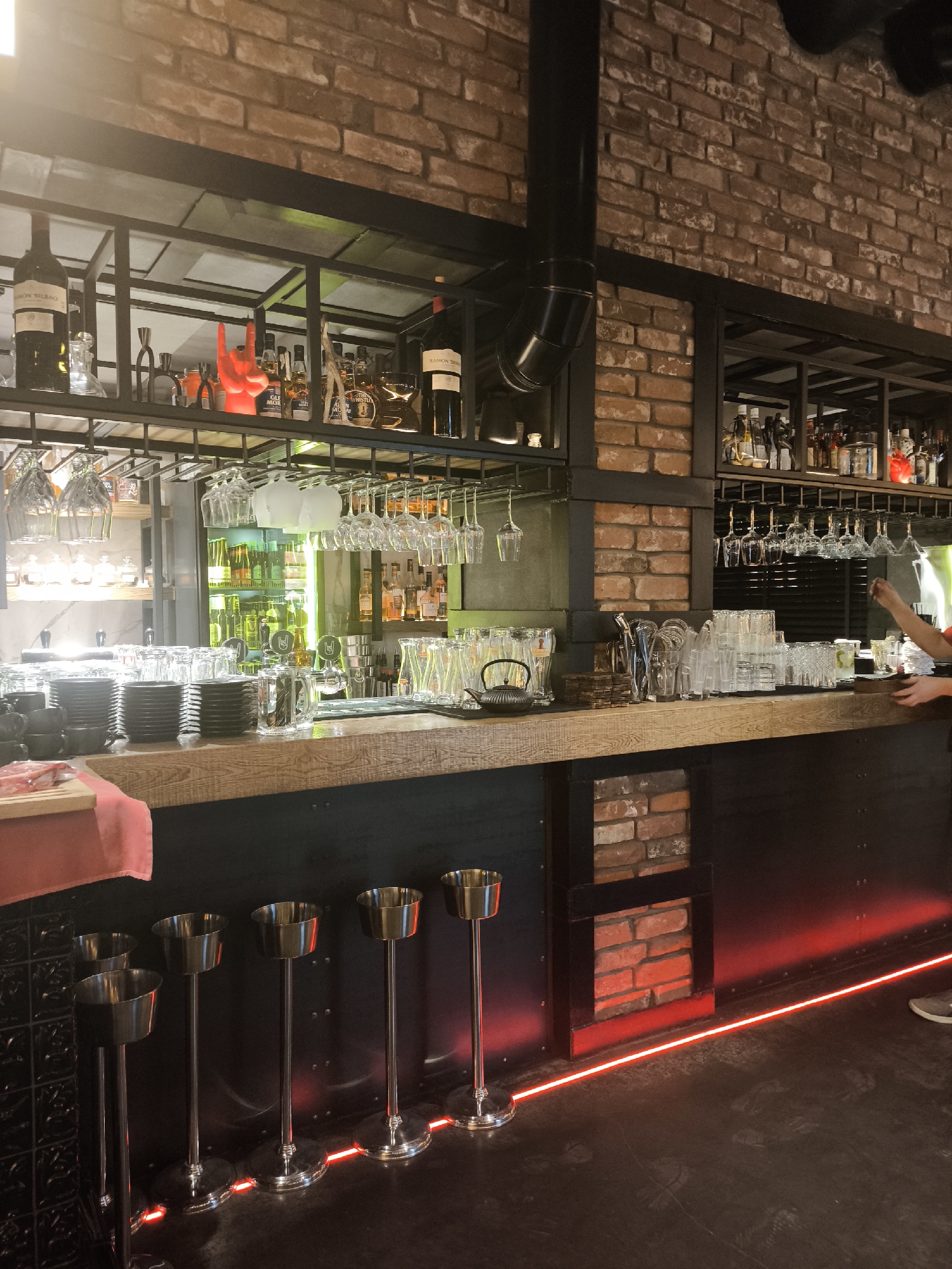 This is an interior shot of a bar.
**Location:** This image appears to be from a location in Izhevsk, Russia, based on visual cues such as potential Cyrillic-scripted labels on items.
**Environment:** The bar features exposed brick walls and a black metal framework around shelving and decorative elements. The lighting includes overhead illumination as well as a red LED strip lighting along the base of the bar.
**Objects:**
* Wine and liquor bottles are arranged on shelves behind the bar.
* Various glassware, including wine glasses and beer mugs, are hanging above the bar or stacked on the counter.
* A kettle is visible on the bar.
* Several ice buckets are positioned along the front of the bar.
* The bar counter itself has a wooden top.
* Black metal and brick paneling is visible on the lower portion of the bar.
**People:**
* A person's arm is visible behind the bar, possibly in the act of serving a customer.