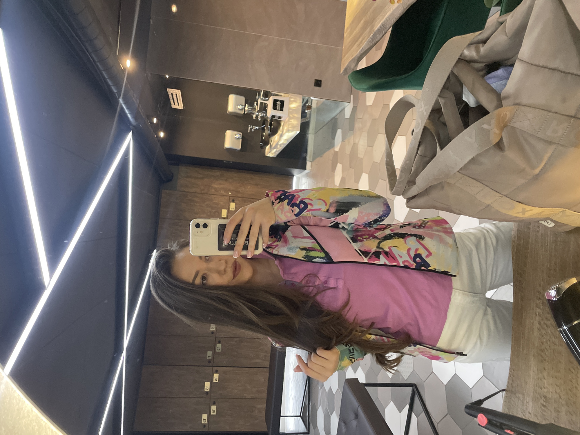 A young woman is captured in a mirror selfie, standing in what appears to be a modern indoor space. She has long, dark brown hair and is wearing a pink collared shirt, white pants, and a vibrant, multi-colored jacket with an abstract pattern. She holds a light-colored smartphone to take the picture, with a subtle smile on her face.
The setting features dark grey/black walls adorned with striking white linear LED lights arranged in geometric patterns. In the background, visible through the mirror's reflection, are several elements: a section of wall with grey and white hexagonal tiles, and a series of dark wooden lockers with reversed numbers (J1 through J6 are partially visible). A dark countertop or shelf runs across the foreground, with a black hairdryer-like object and part of a green armchair also reflected. A large, beige quilted bag with wide straps, possibly bearing the text "J.OXA" or similar, hangs behind her. Overhead, some ceiling fixtures and additional small lights are visible. The overall ambiance suggests a contemporary locker room, gym changing area, or stylish public space. The reflection of her phone screen shows partial, reversed text that looks like "B L L I N G."