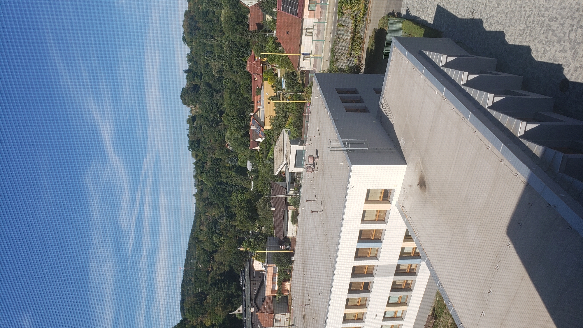 The image shows a view from a building, looking out over a residential area towards a tree-covered hillside under a clear blue sky with wispy clouds. In the foreground, there are two flat rooftops. The closer rooftop is partially in shadow and appears to be made of a light-colored material with a grid pattern, possibly a safety net. It has several metal fixtures and a dark stain. The further rooftop is also flat and light-colored, with a prominent white building extending from its edge. This building has multiple windows with light-colored frames and some blue accents. The rooftop of this building has several ventilation pipes and a ladder.
Beyond the rooftops, a neighborhood of houses with red and orange tiled roofs is visible. The houses are surrounded by trees and greenery. In the distance, a forested hill rises, with some smaller structures and possibly a communication tower on its summit. The overall impression is of a sunny day in a suburban or semi-rural environment.
Based on visual cues and common architectural styles, this scene is likely located in Prešov, Slovakia, a city known for its hilly terrain and mixture of modern and older residential buildings. The specific location might be near the edge of the city, bordering a natural area. There are no people visible in the image. The scene depicts a static environment, with no apparent activity or interaction.
