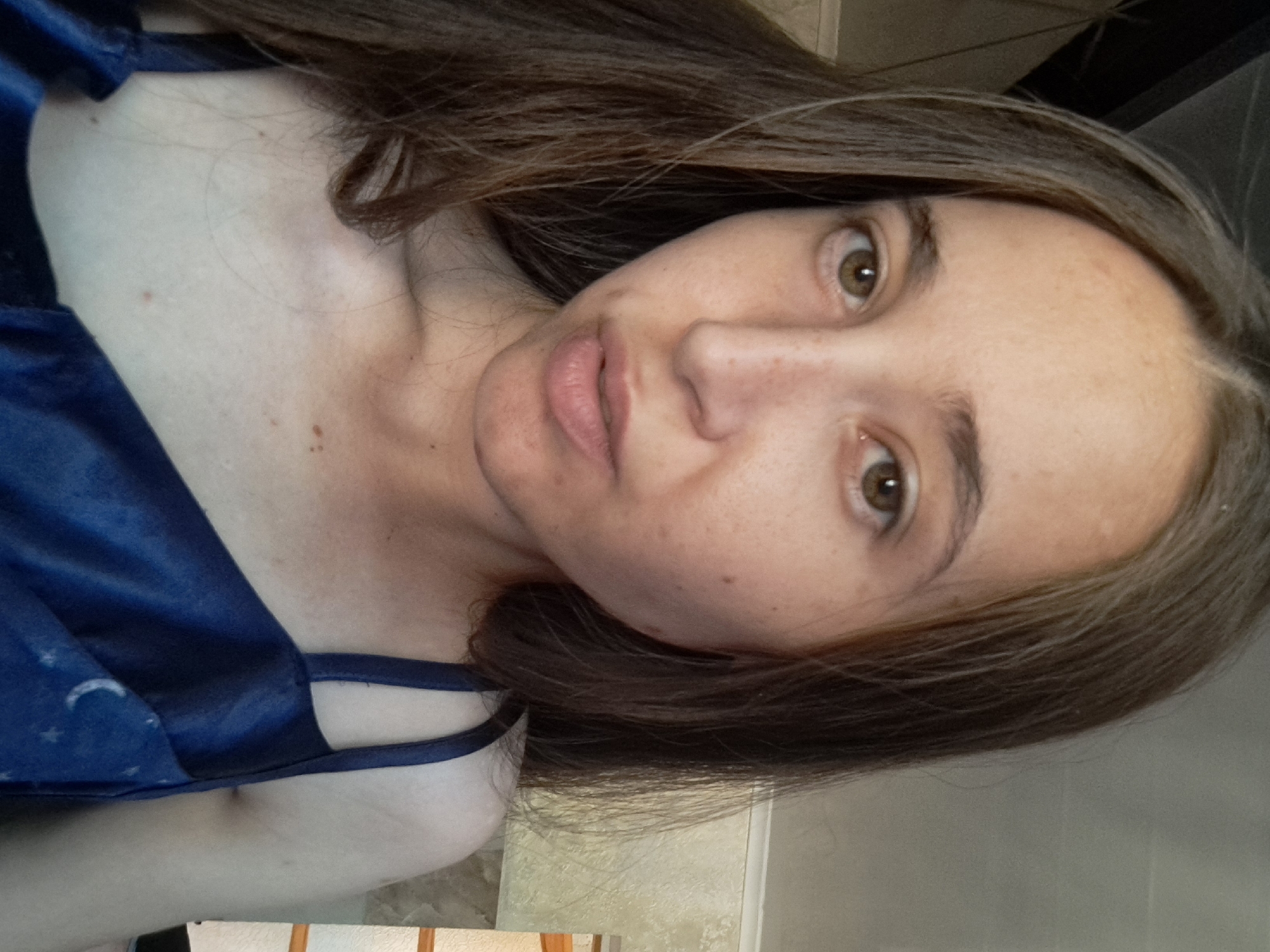 A close-up, selfie-style photograph features a young woman with long, brown hair. Her face is angled slightly to the right, and she has light brown eyes and freckles across her nose and cheeks. She is wearing a dark blue satin top with thin straps, adorned with a celestial pattern of stars and moons. The background appears to be an indoor setting, possibly a bedroom, with a neutral-toned wall and a sliver of what might be a wooden structure on the right side of the frame. The lighting suggests it is daytime. No specific location within Vypasne, Ukraine, can be identified from the image.