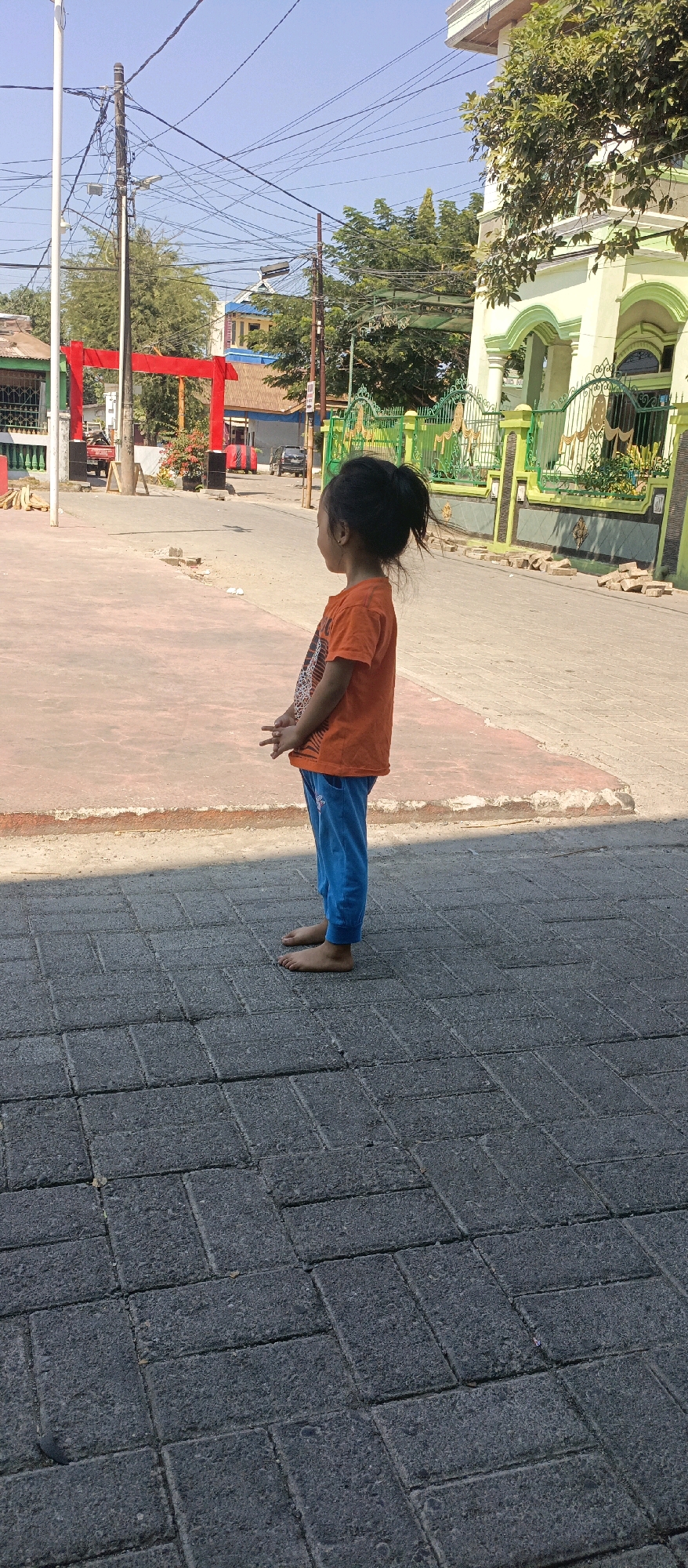 A young girl, wearing an orange t-shirt and blue pants, stands on a brick-paved sidewalk in Makassar, Indonesia. She is facing away from the camera, looking to her right. Her hands are raised slightly in front of her, with her fingers extended. In the background, a street is visible, lined with a green fence and a building. A red archway is also present in the distance. The sky is clear and blue, and the sun is shining, casting shadows on the ground. The environment appears to be an urban or suburban setting.