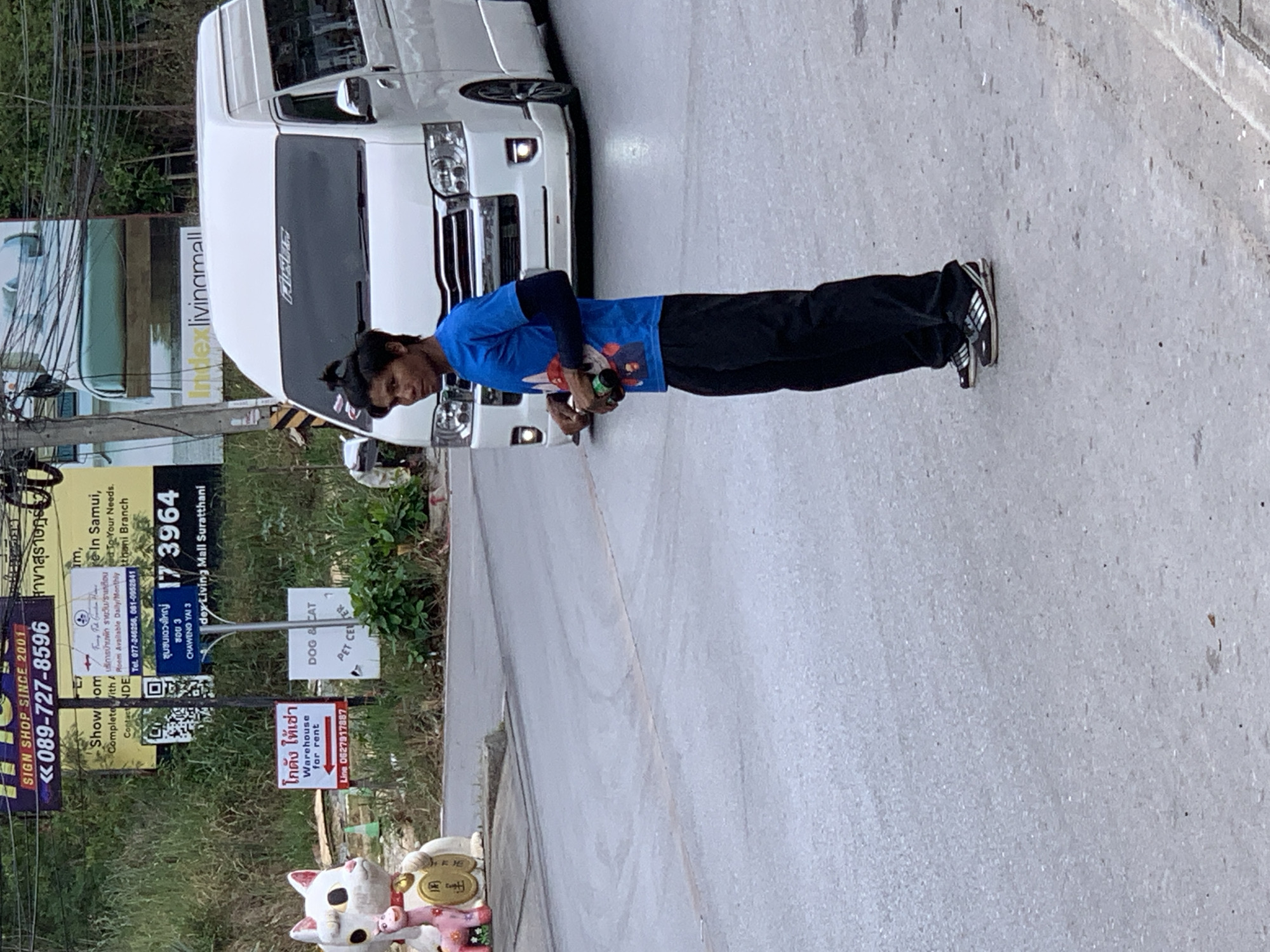 A young Southeast Asian male stands on a paved road, facing slightly left, with a white van behind him. He wears a blue t-shirt with a graphic design and black pants. His hair is pulled up in a bun. He holds what appears to be two cans in his right hand. The van has tinted windows and a sticker with a logo on the windshield. In the background, to the left of the man, is a large white cat statue with a pink figure beside it. Several signs are visible on poles: one in Thai and English advertising "SIGN SHOP SINCE 2001" with a phone number, another with a QR code, and a sign that reads "DOG & CAT PET CENTER". Further in the background, large billboards with text in Thai and English are partially visible. The scene appears to be in a commercial area.