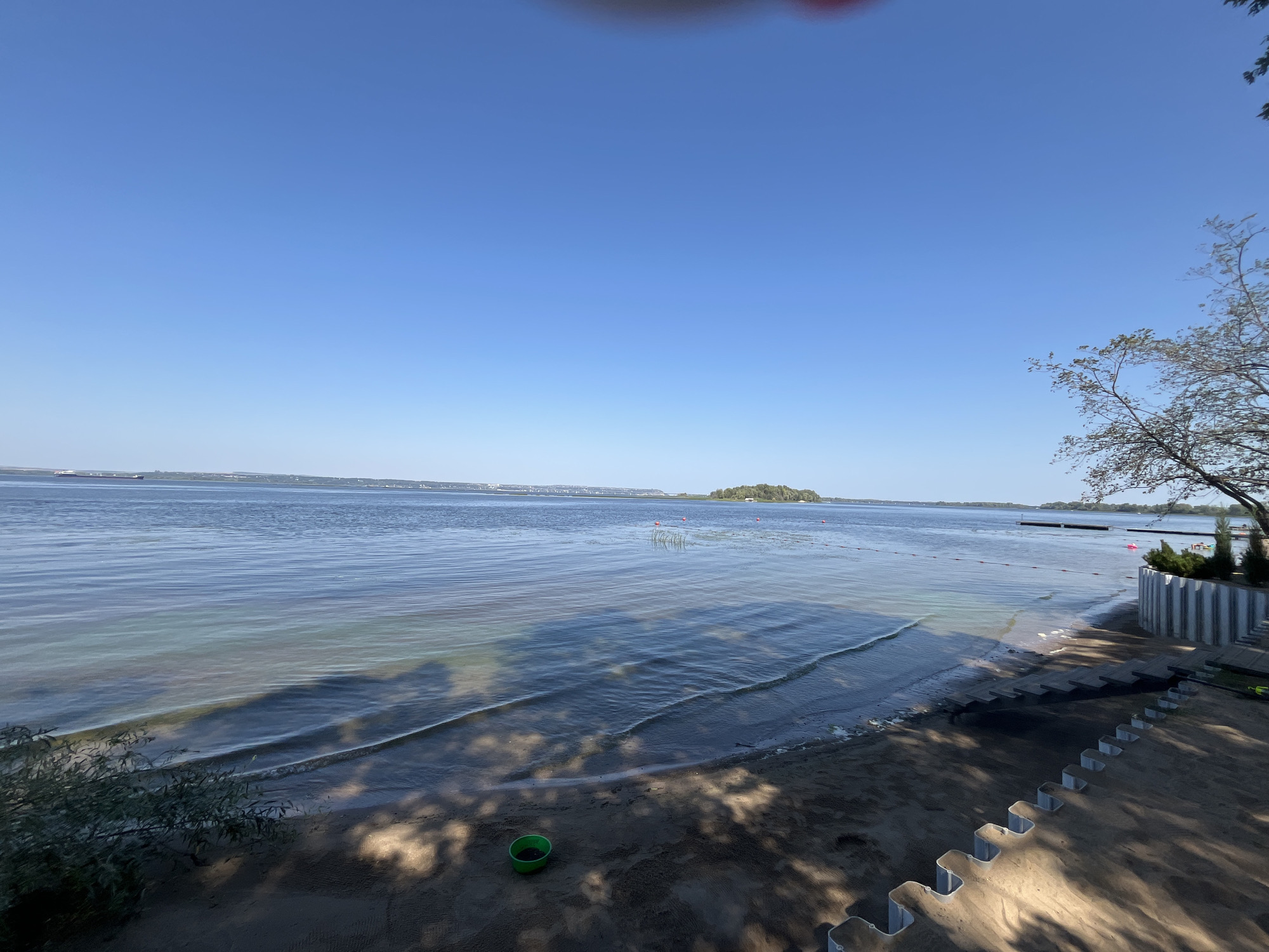 The image shows a sandy beach next to a calm body of water under a clear blue sky. In the distance, a small, tree-covered island is visible, and a large ship can be seen on the horizon. The beach has a modern, stepped barrier made of concrete or stone. To the right, there are some manicured bushes and a tree with visible branches reaching into the frame. The water's edge is gently lapping onto the sand, and there are some red buoys floating in the water, possibly marking a swimming area. The scene is illuminated by bright sunlight, casting shadows from the tree onto the sand. There is a green bucket on the sand near the water's edge. The image does not provide specific visual cues to pinpoint its exact location within Shumeika, Russia, but it depicts a typical waterfront recreational area.