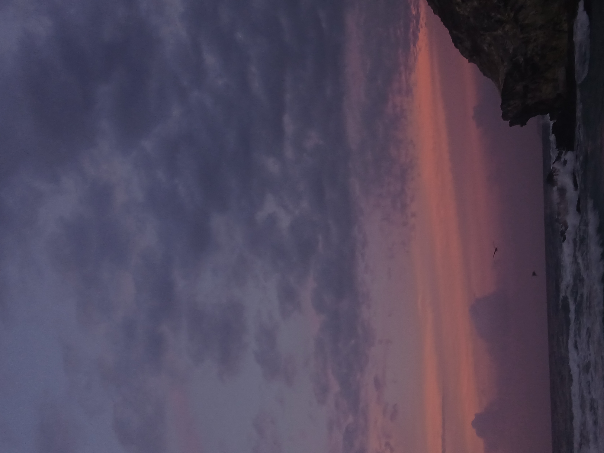A vertical shot shows a dark, cloudy sky above the ocean at Playa Parguito, Venezuela. The sky has hues of purple, gray, and pink. In the upper right corner, there is a rocky cliff. The ocean, a dark gray, appears to be turbulent with white foamy waves.