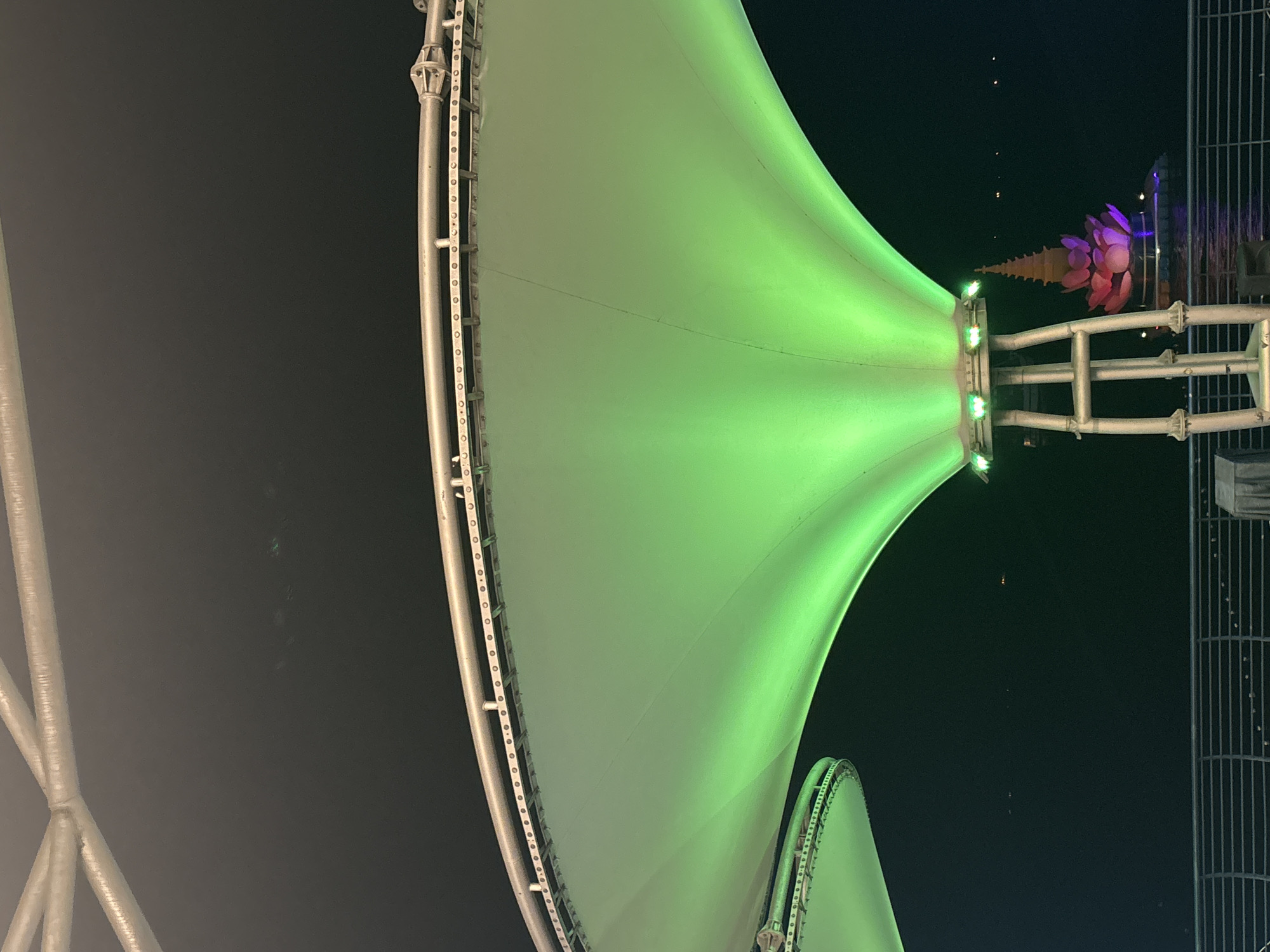 The image depicts a nighttime scene in North Jakarta, Indonesia, featuring a large, green-lit fabric canopy structure. The canopy curves downwards, supported by metal scaffolding. Below the canopy, in the distance, a lotus-shaped structure illuminated with pink and purple lights stands on the water. A metal railing runs across the foreground, suggesting an elevated walkway or viewing platform. The sky is dark, with a few faint lights visible in the distance, possibly from boats or other structures. The overall impression is of a modern waterfront development.