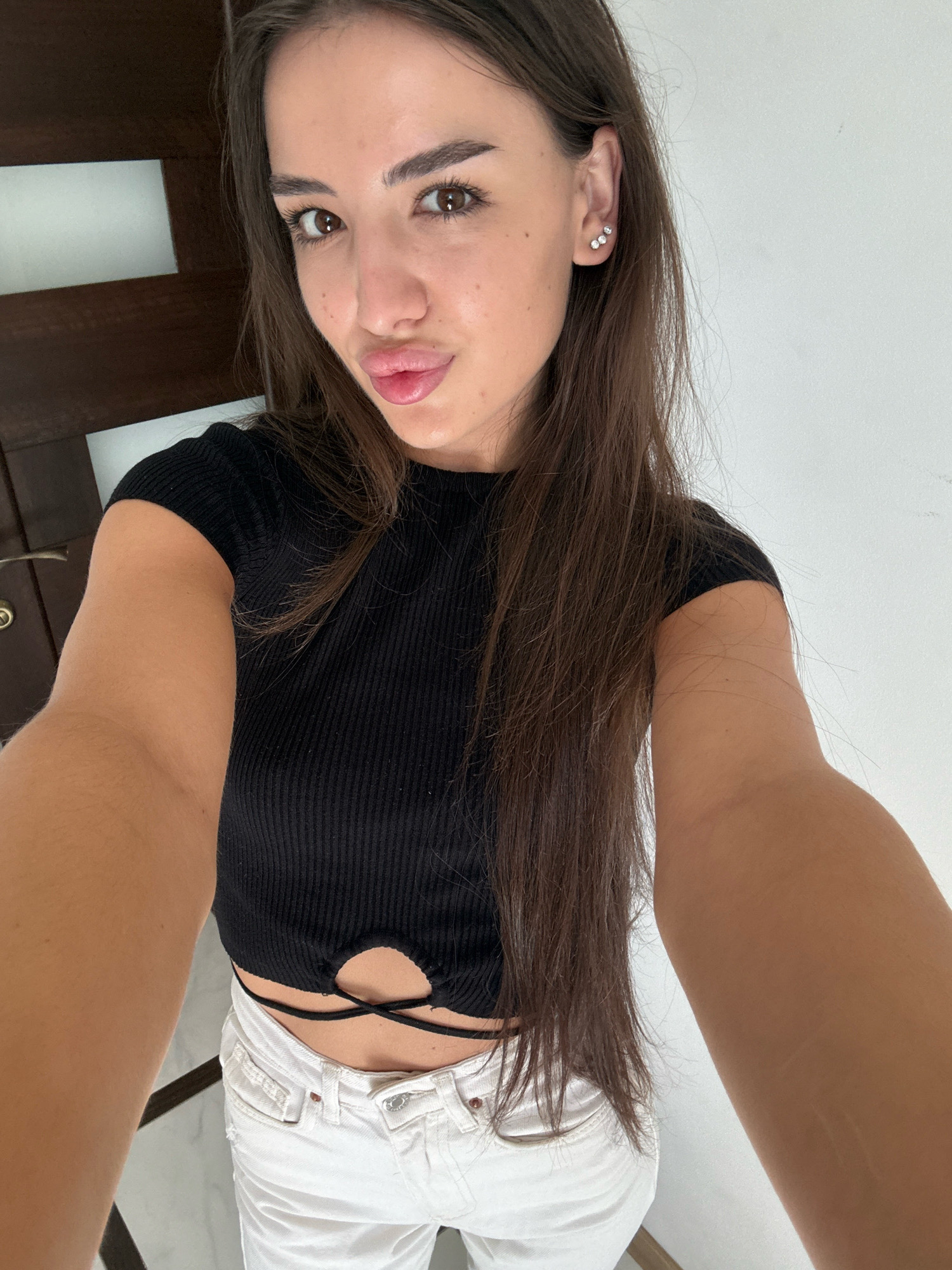A young woman with long brown hair and brown eyes is taking a selfie. She is wearing a black ribbed crop top and white jeans. She has on a stud earring with three small stones on her left ear. The background appears to be an indoor setting with wooden elements and a white wall. There is no visible indication of the location being Chișinău, Moldova.