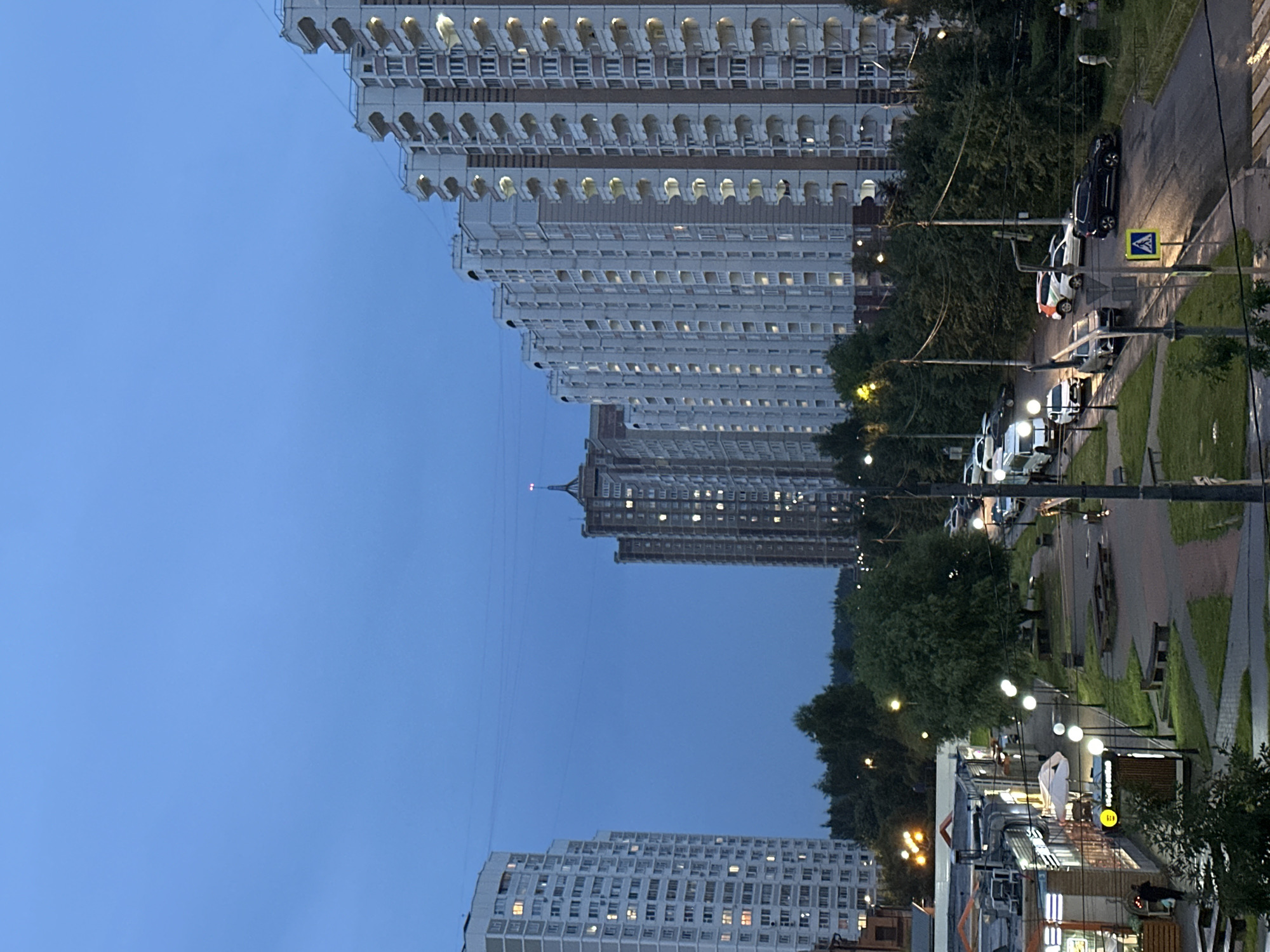The image depicts an evening scene in the city of Podolsk, Russia, with a twilight sky overhead. Dominating the right side of the frame are several tall, multi-story residential buildings with cream-colored facades and numerous balconies. To the left, another tall residential building, predominantly white, stands partially visible. In the mid-ground, a street is lined with trees and streetlights, with several cars parked and driving. A low-rise commercial building with illuminated storefronts is visible on the far left. The ground appears wet, suggesting recent rain. In the foreground, a paved area with benches and planters suggests a public space. The overall impression is of a residential urban environment transitioning into evening.