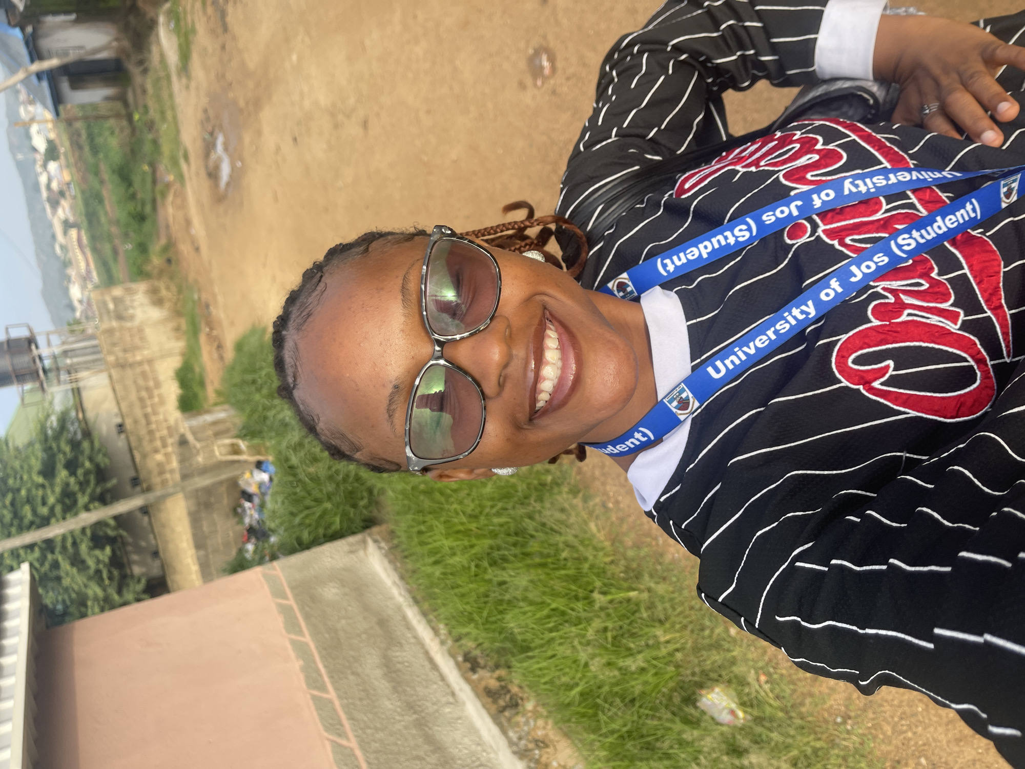 A medium shot selfie shows a Black woman smiling, wearing sunglasses, a black and white striped shirt with "Original" in red script on the front, and a blue lanyard that says "University of Jos (Student)". She is standing outdoors with a dirt road behind her, and a building and green grass to her left. The background shows a distant town or village with houses and hills, suggesting a location in the city of Rafin Par, Nigeria.