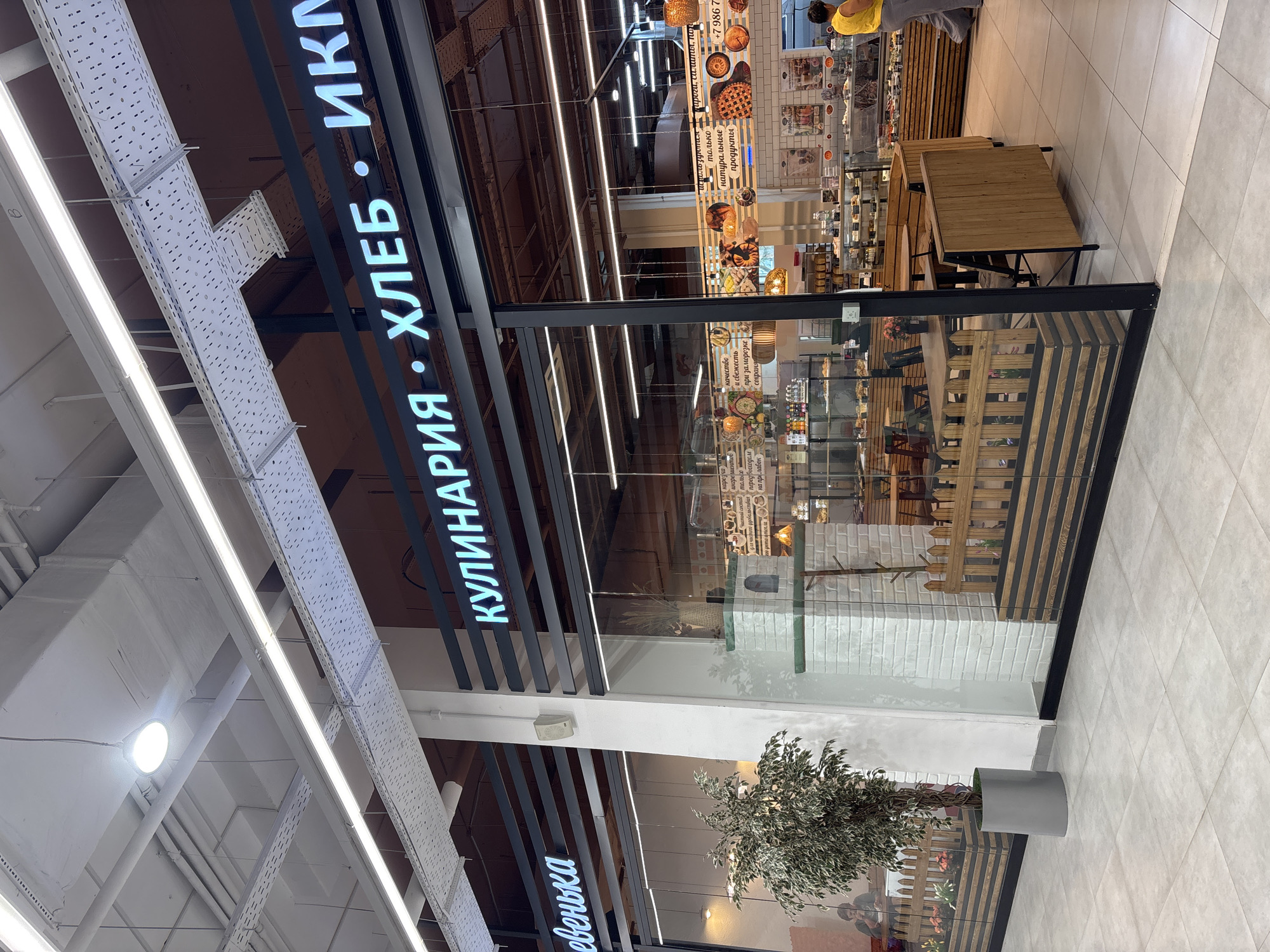 The image shows the exterior of a bakery and cafe with a glass storefront. The name of the establishment is displayed in large, illuminated letters. On the left side of the storefront, the word "Евенька" is visible. To the right, the words "КУЛИНАРИЯ • ХЛЕБ • ИКМ" are displayed. Inside the cafe, tables and chairs are arranged. A person is seated at one of the tables. There is a decorative plant in a grey pot outside the storefront. Further inside the establishment, various baked goods and food items are displayed in refrigerated cases. In the background, a woman in a yellow top and grey pants is walking. The floor is tiled. The overall environment appears to be an indoor shopping area or mall. The location of the bakery is identified as Naberezhnye Chelny, Russia.