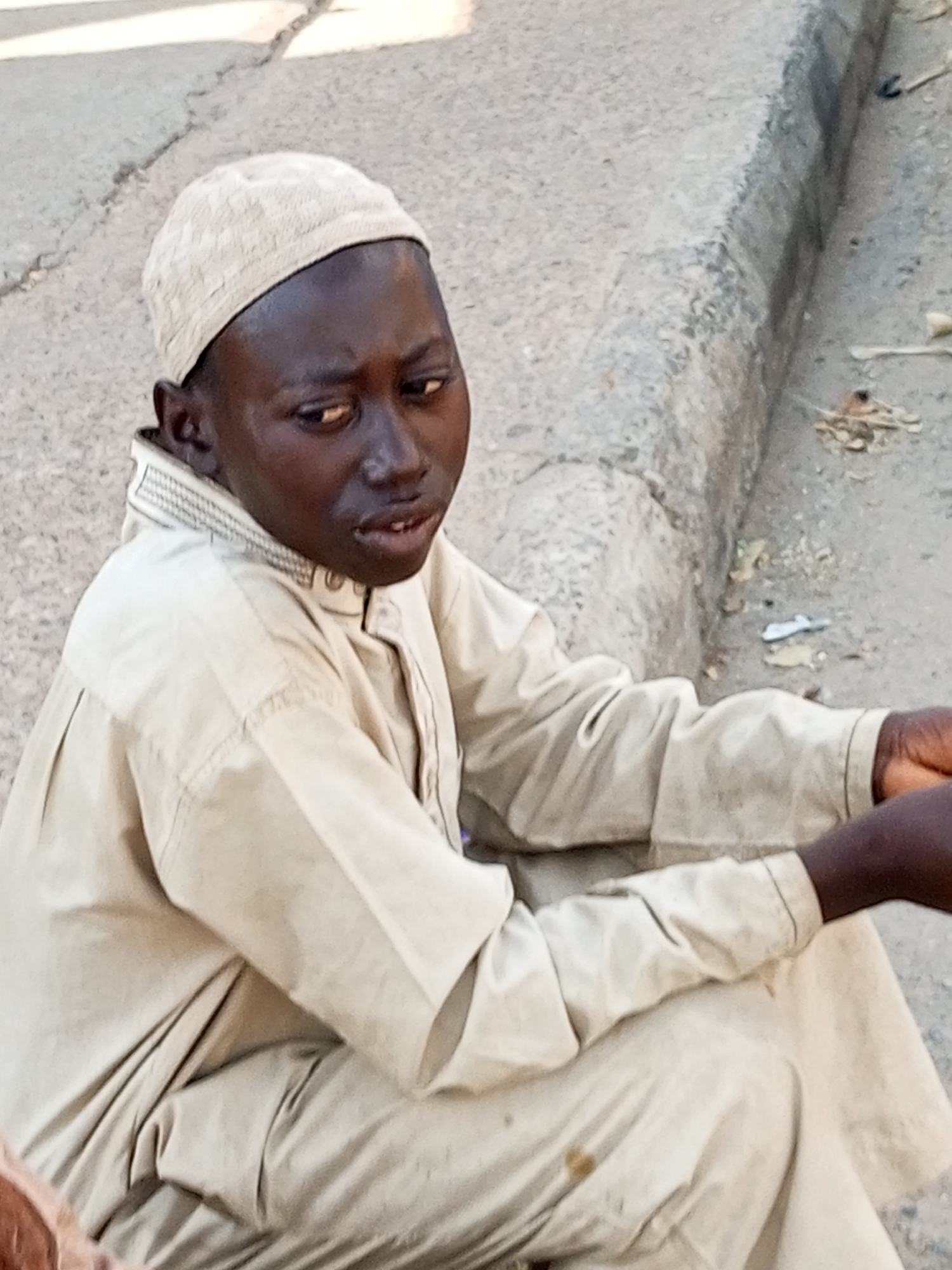 A young Black male, wearing a light-colored cap and a matching light-colored robe, sits on a cracked concrete surface. He is facing the right of the frame, with his body slightly turned towards the viewer. The robe has a collar with visible stitching. His right arm is resting on his leg, and his left arm is not fully visible. His facial expression is ambiguous, with his eyes looking slightly upwards and to the side. The background consists of a curb made of concrete, with some debris scattered on the ground next to it, including small twigs and what appears to be a piece of white plastic. The overall setting suggests an outdoor, possibly urban environment. The image appears to have been taken in Arawa, Nigeria.