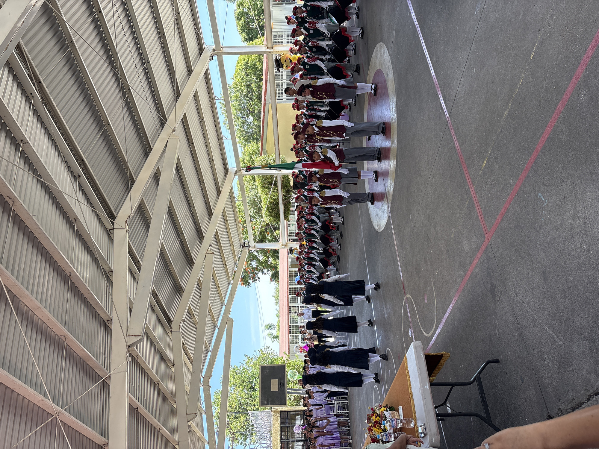 In the outdoor courtyard of a school in Tejalpa, Mexico, a formal ceremony is underway beneath a corrugated metal roof supported by white metal beams. A group of students, dressed in dark uniforms with white accents, are facing away from the camera, appearing to be part of a performance or procession. To their right, a marching band or drum corps, also composed of students in uniforms with red hats, stands at attention, some holding drums. A Mexican flag is prominently displayed, held by two students. In the background, other students and adults are gathered, and a basketball hoop is visible. A table set up for an event is in the foreground, adorned with flowers and water bottles. The ground is a concrete court marked with colored lines. The overall scene suggests a school assembly, award ceremony, or patriotic event.