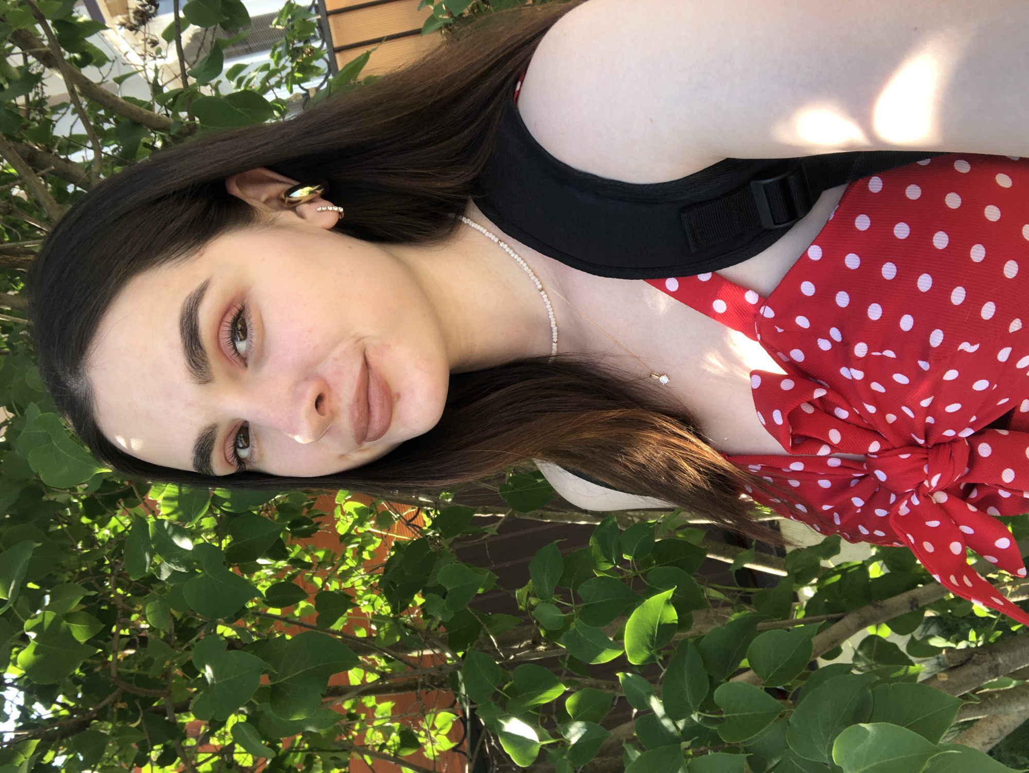 A woman is captured in a medium shot, her face angled towards the camera. She has long, dark brown hair and fair skin. She is wearing gold earrings and a white beaded necklace with a small gold pendant. Her outfit is a red dress with white polka dots. A black strap, possibly from a backpack, is visible over her shoulder. The background shows green foliage, suggesting an outdoor location. The light source appears to be natural sunlight. There is no indication that this picture was taken in Noviy, Russia.