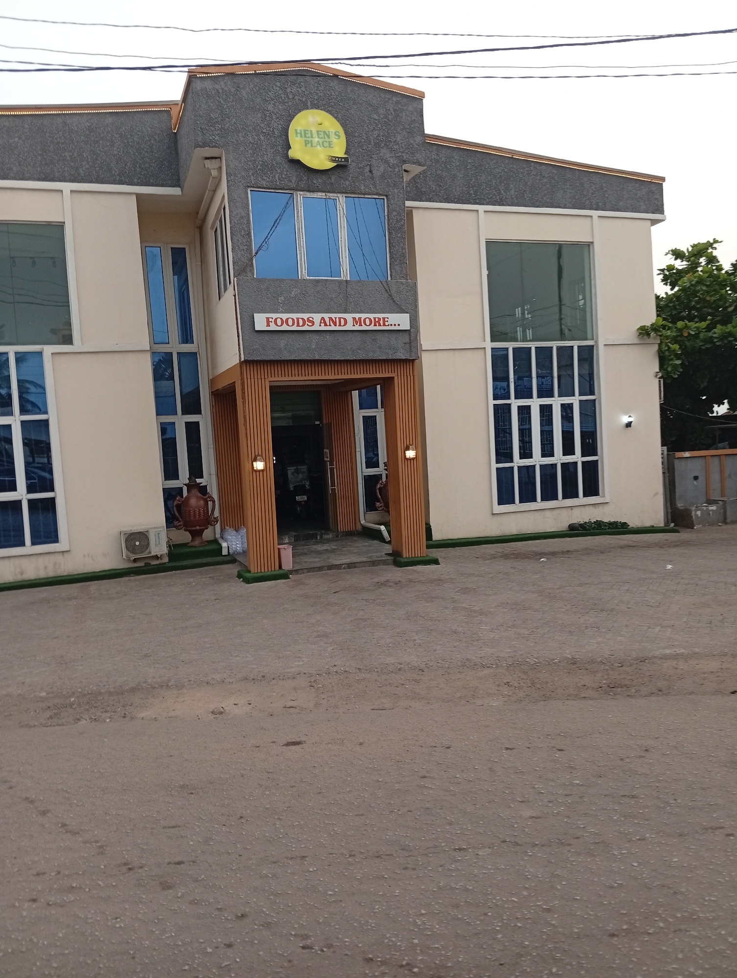This full-shot image captures the exterior of "Helen's Place," a commercial establishment in Lokoja, Nigeria, under what appears to be an overcast sky or late afternoon lighting. The two-story building features a modern design with light cream-colored walls, large blue-tinted windows, and dark grey textured accents on the upper facade.
Prominently displayed on the front of the building is a circular yellow sign with the text "HELEN'S PLACE" in black, with "SUPER" or "SUPPER" visible in smaller text beneath. Below this sign, above the main entrance, another sign reads "FOODS AND MORE...". The entrance itself is recessed and framed by vertical wooden slats, with wall-mounted lights on either side.
To the left of the entrance, an air conditioning unit is mounted on the wall, and a large, brown, decorative urn or vase sits on a patch of artificial green turf. Similar green turf lines the base of the building along the front.
The foreground consists of a wide, unpaved or lightly paved ground area, suggesting a parking lot or access road, which appears somewhat dusty. Overhead, several power lines stretch across the visible sky. A tree with green foliage is partially visible on the far right, bordering the property. No people or overt activities are visible, presenting a serene, almost deserted, commercial scene.