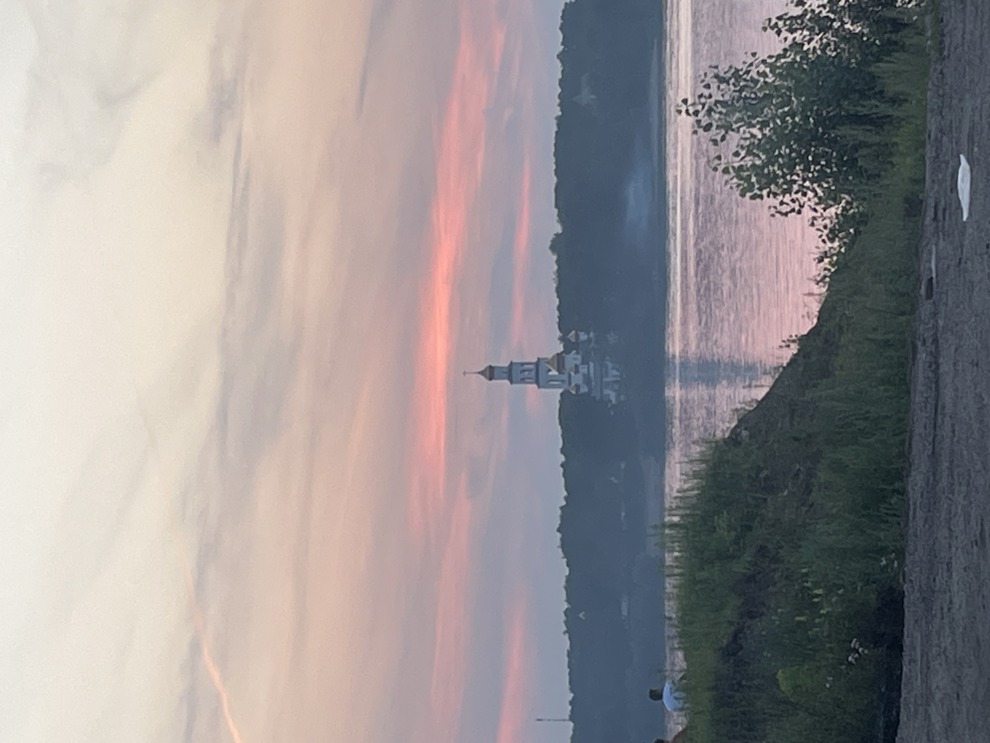 The image shows a wide shot of a river at dusk, with a church on the opposite bank. The sky is filled with soft pink and orange hues, indicating the sunset. The river's surface reflects the colors of the sky, creating a serene atmosphere. On the foreground bank, there is a grassy slope with some people sitting or standing. The church, a prominent structure with a tall white tower and golden domes, is silhouetted against the vibrant sky. The scene conveys a sense of peace and tranquility, capturing the beauty of nature and architecture at twilight.