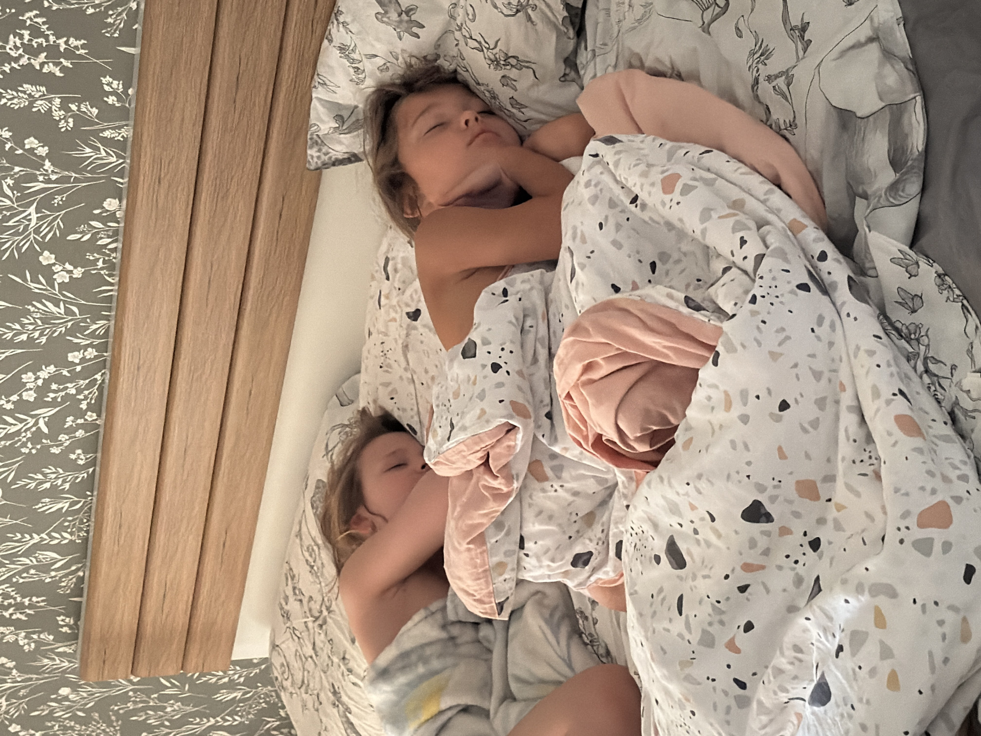 Two children are sleeping in a bed. One child is on the top, curled up with their arms around their head. The other child is below, also curled up. They are covered with a patterned blanket and a pink blanket. The bed has patterned sheets. There is a wooden headboard next to a wall with patterned wallpaper. The location is not discernible from the image.
