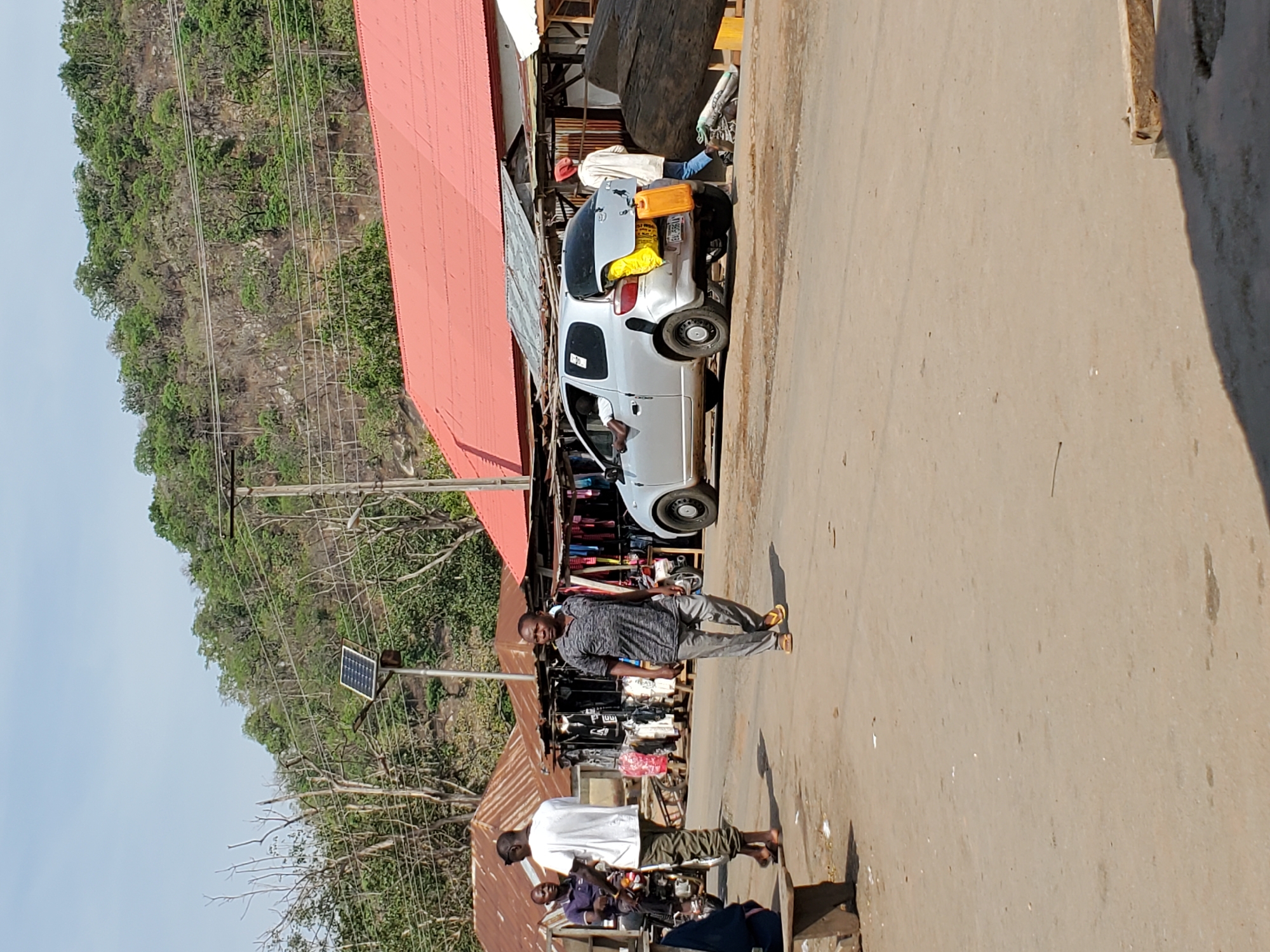The image captures a street scene in Mararaba, Nigeria, under bright daylight conditions. The paved road occupies the central and lower portions of the frame.
Along the left side of the road, a row of market stalls and buildings with distinctive red corrugated iron roofs are visible. These stalls appear to be selling various goods, including clothing. A silver hatchback car is parked facing right with its trunk open, containing yellow bags and an orange jerrycan, suggesting loading or unloading activity. A person in a white garment and blue pants is partially visible near the car.
Several people are present on the street. A man in a dark grey patterned shirt, light grey pants, and yellow sandals walks towards the viewer in the foreground. Further along the road, another man wearing a white t-shirt and camouflage pants stands near a motorcycle, close to a market stall. A third person in a dark blue shirt is partially visible near the second man.
The background features a steep, densely vegetated hillside covered in green trees, indicating a natural landscape bordering the commercial area. Utility poles and a network of power lines traverse the scene. A solar panel is mounted on a pole, visible towards the middle right.
The bright sunlight and clear sky indicate it is daytime. The license plate on the silver car is partially visible and reads "KYA-66ETB". The scene portrays everyday life and commerce in a semi-urban setting, with a mix of natural and built environments.