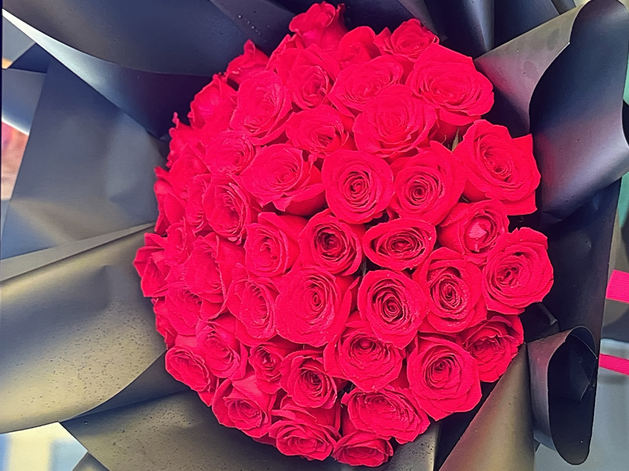A large, vibrant bouquet of red roses is prominently featured, tightly arranged and filling the central part of the frame. The roses are a deep, rich crimson, with numerous individual blooms densely packed together. Each rose displays intricate layers of soft, velvety petals, some still partially closed, others more open.
The bouquet is elegantly wrapped in black paper, which contrasts sharply with the bright red flowers. The wrapping paper has a matte finish and appears to be made of a textured, possibly slightly crinkled material, folded neatly around the base and sides of the floral arrangement. The folds are distinct, creating a structured and sophisticated presentation.
The setting appears to be indoors, with a dark, indistinct background that ensures the entire focus remains on the bouquet. No people are visible, and no specific activity is taking place beyond the static display of the prepared flowers. The lighting is even, suggesting an artificially lit environment.
The overall impression is one of beauty, romance, and luxury. There is no visible text in the image, nor are there any specific location cues to identify the setting as Shumen, Bulgaria, or any other particular place. The bouquet is a classic symbol of love and affection.