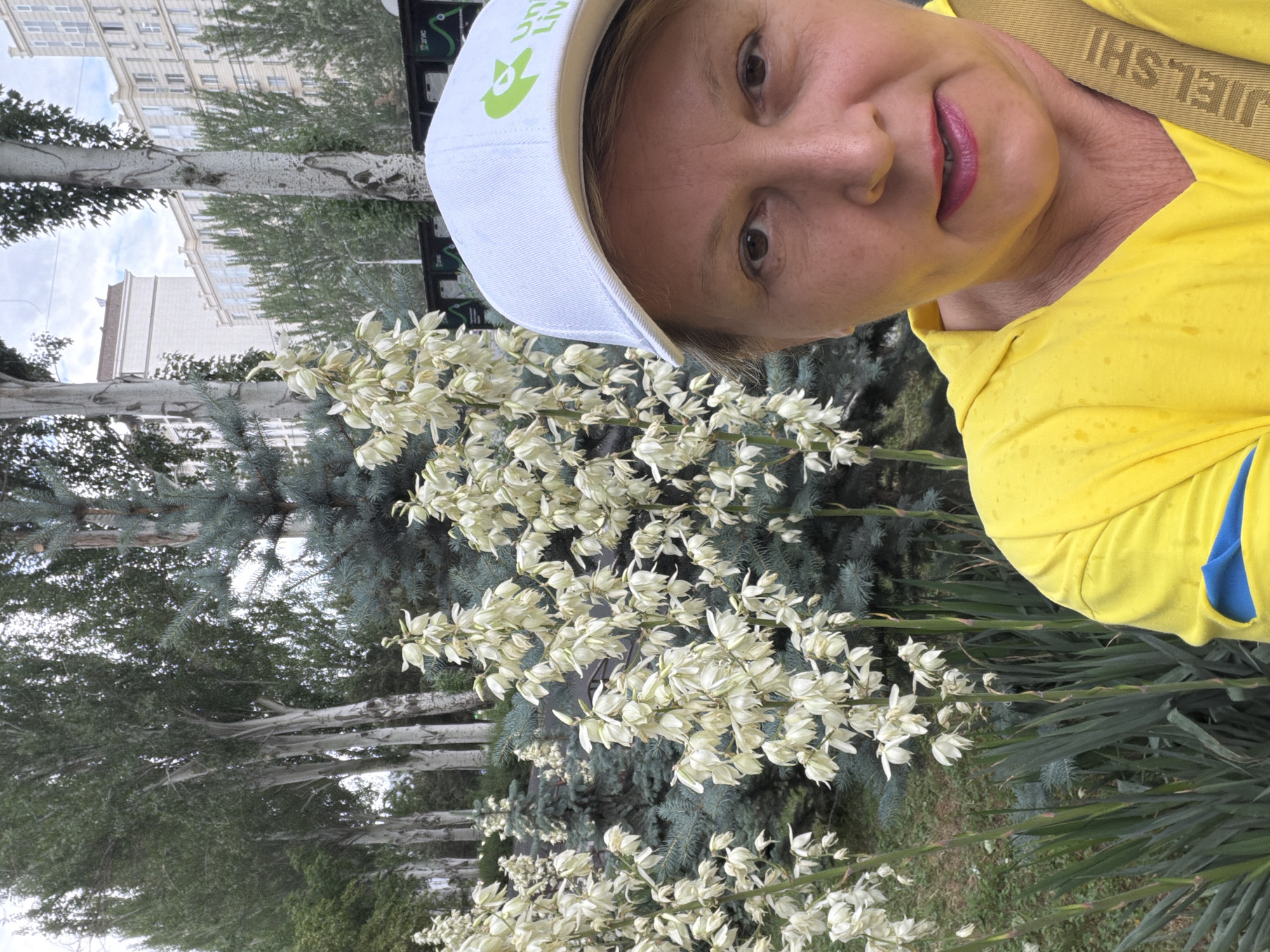 A woman in a yellow hooded jacket and a white cap with a green logo is taking a selfie outdoors. She is positioned on the right side of the frame, with her face and upper chest visible. The woman has blonde hair, brown eyes, and is wearing lipstick. The cap has the words "UJIELSHI" printed on the strap. Behind her, in the background, are tall trees with white flowers and blue-green coniferous trees. The environment appears to be an urban setting, with buildings visible in the distance. The lighting suggests it is daytime.