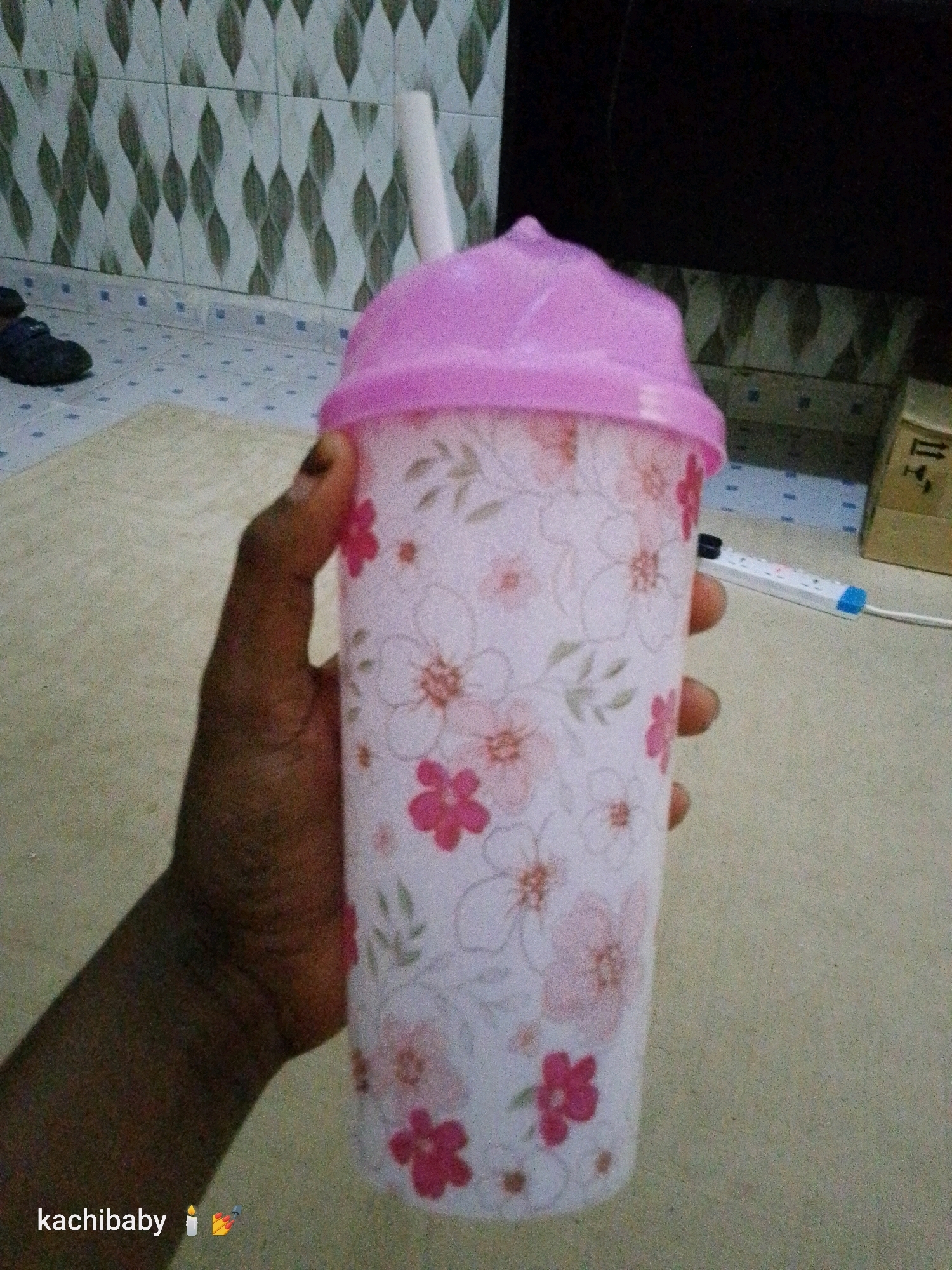 A hand holds a plastic tumbler with a pink lid and a white straw. The tumbler has a floral pattern of pink and white flowers. The background shows a tiled wall and a light-colored floor. A power strip is visible on the floor in the background. The image is a close-up of the tumbler and hand, with the background out of focus. The scene appears to be indoors.