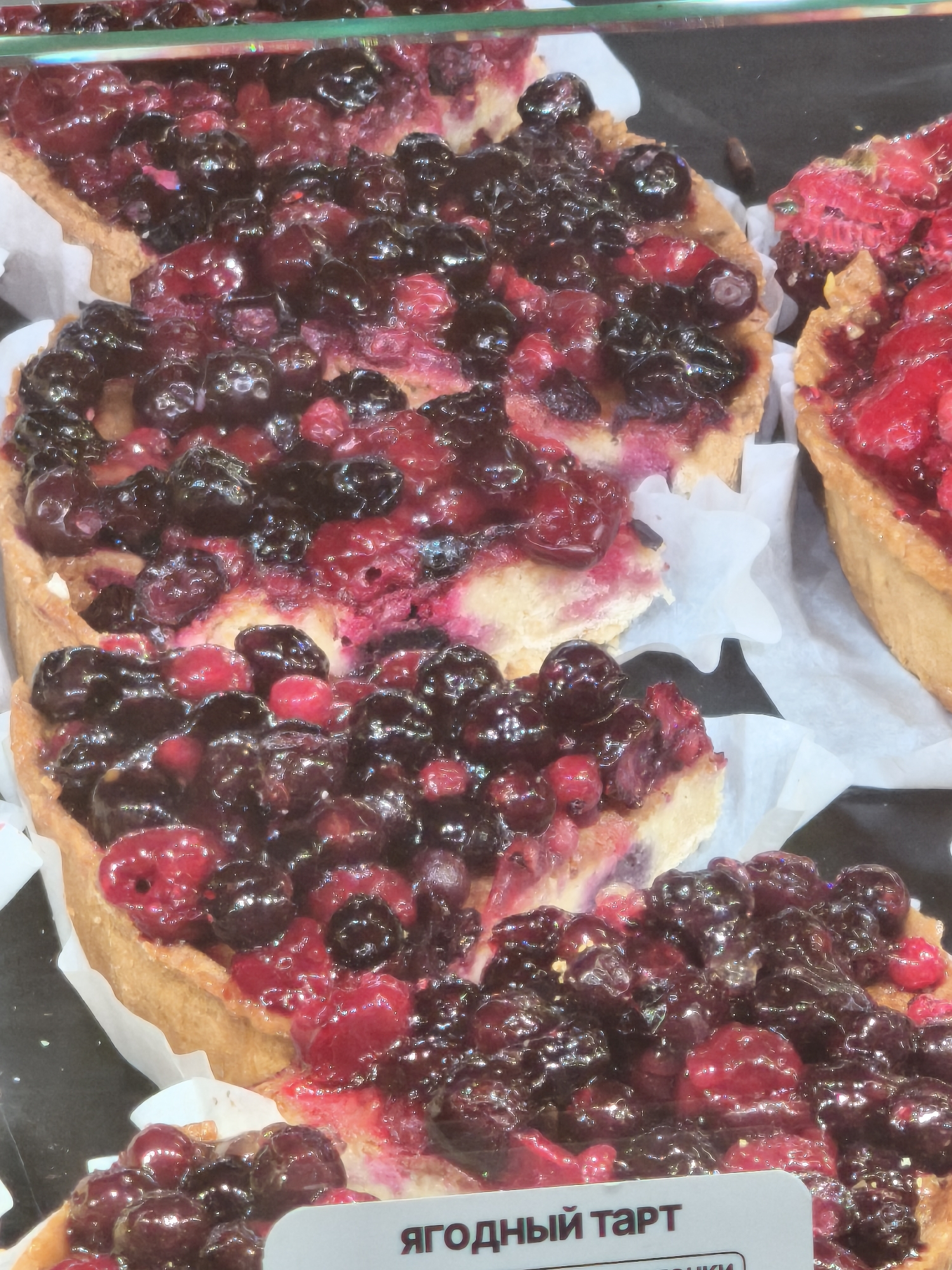 Two berry tarts are displayed on a black surface, partially covered by white paper liners. The tart on the left is filled with blueberries and raspberries, with a golden-brown crust. The tart on the right is filled with raspberries and appears to have a similar crust. A white label with Russian text is partially visible at the bottom of the image. The overall lighting is bright, with reflections visible on the glossy surface of the berries.