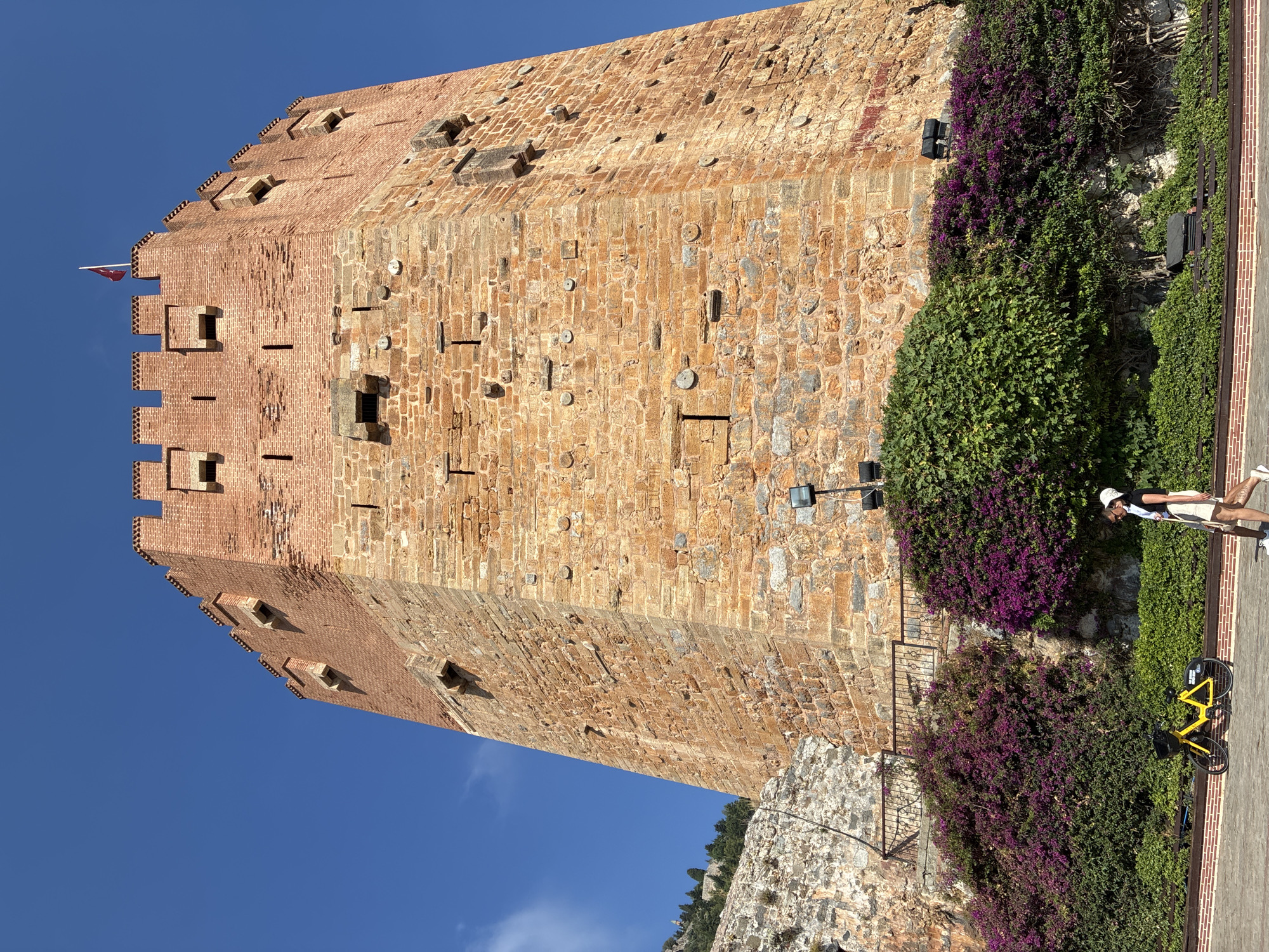 The image depicts the historic Kızıl Kule (Red Tower) in Alanya, Turkey, a prominent landmark and a symbol of the city. The tower, constructed of red brick and stone, stands tall against a clear blue sky. Its weathered facade displays architectural details such as crenellations at the top and numerous small rectangular openings.
In the foreground, lush green bushes and vibrant purple bougainvillea flowers form a dense border along the base of the tower. A yellow bicycle with a black basket is parked on the paved walkway. Two people are walking from left to right in the lower right portion of the frame. The individual closer to the viewer is a person wearing a white cap, a dark top, and light-colored shorts, carrying a bag. The second person is partially obscured. The ground in front of them is paved with brickwork.
The overall scene captures a sunny day with a mix of historical architecture, natural elements, and everyday life in Alanya.