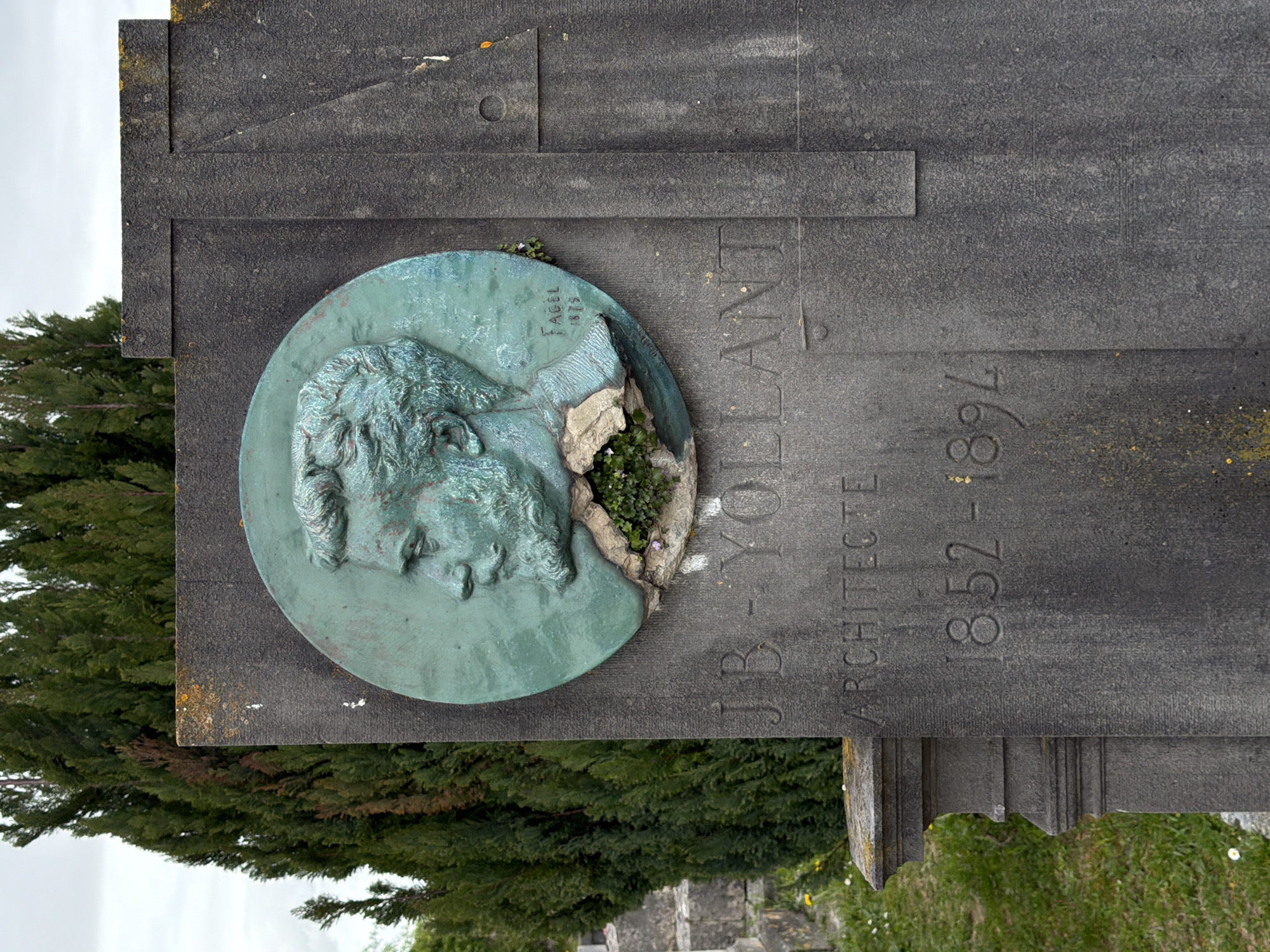 This image depicts the gravestone of J.B. Yollant, an architect, located in Auvers-sur-Oise, France. The gravestone is made of grey stone and includes a bronze medallion featuring a profile of Yollant. The medallion is partially broken, with small plants growing in the damaged section. Text carved into the stone reads "J.B. YOLLANT ARCHITECTE" and "1852-1894". The signature "Fagel 1875" is inscribed on the medallion. A dense evergreen tree or shrub is visible in the background to the left of the gravestone. The sky is overcast. Other graves and vegetation are vaguely visible in the background. The orientation of the image is rotated 90 degrees counter-clockwise.