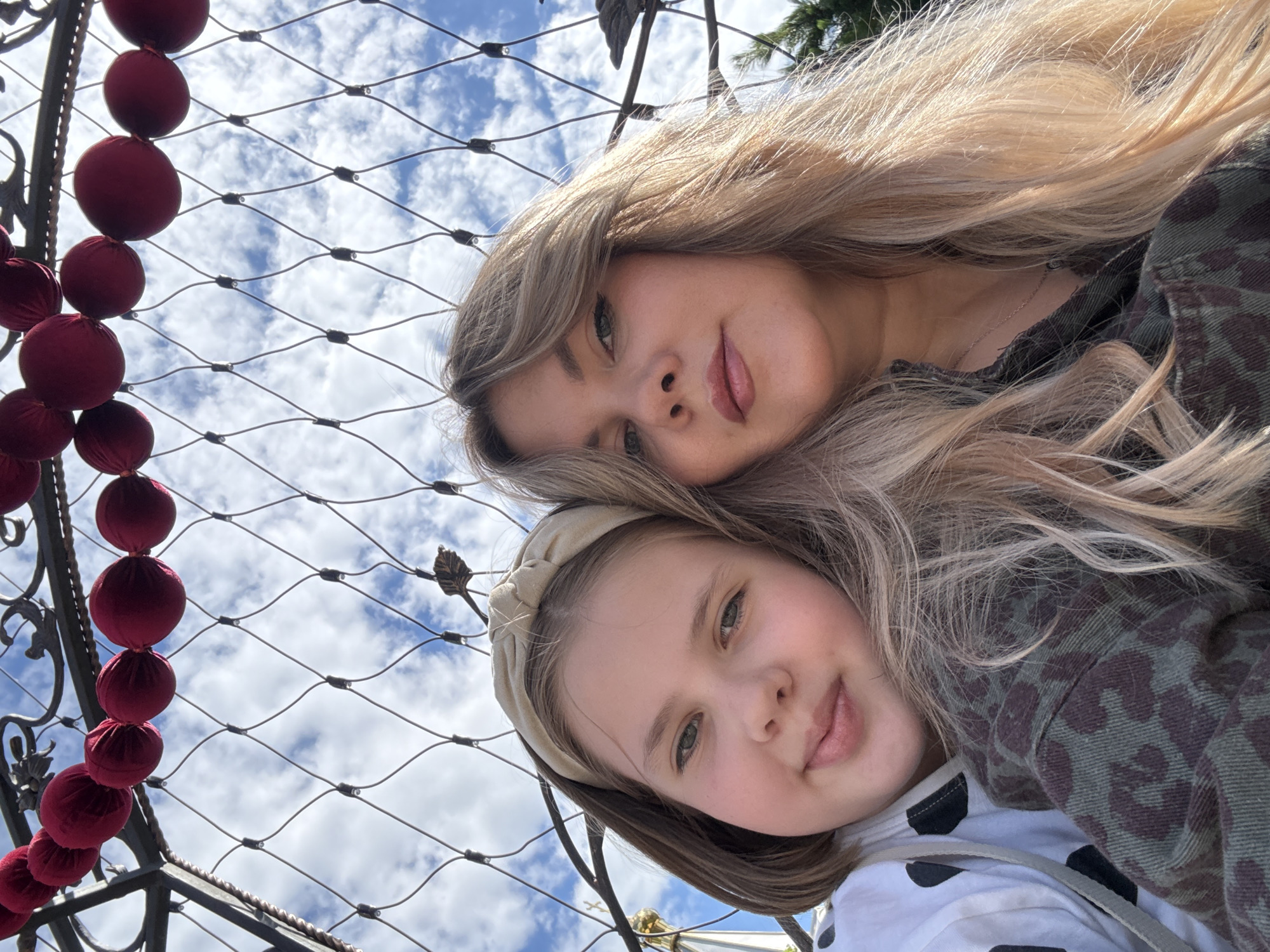 In Izhevsk, Russia, two females are captured in a selfie outdoors. A younger female with shoulder-length brown hair, wearing a beige headband and a white hoodie with black polka dots, smiles gently towards the camera. Beside her, an adult female with long, wavy blonde hair and makeup, wears a green camouflage jacket. They are positioned in front of a wire mesh fence, with a string of red spherical ornaments visible above them against a backdrop of a cloudy blue sky. The background hints at an outdoor location, possibly a park or an area with decorative elements, with a glimpse of architectural structures in the distance.
