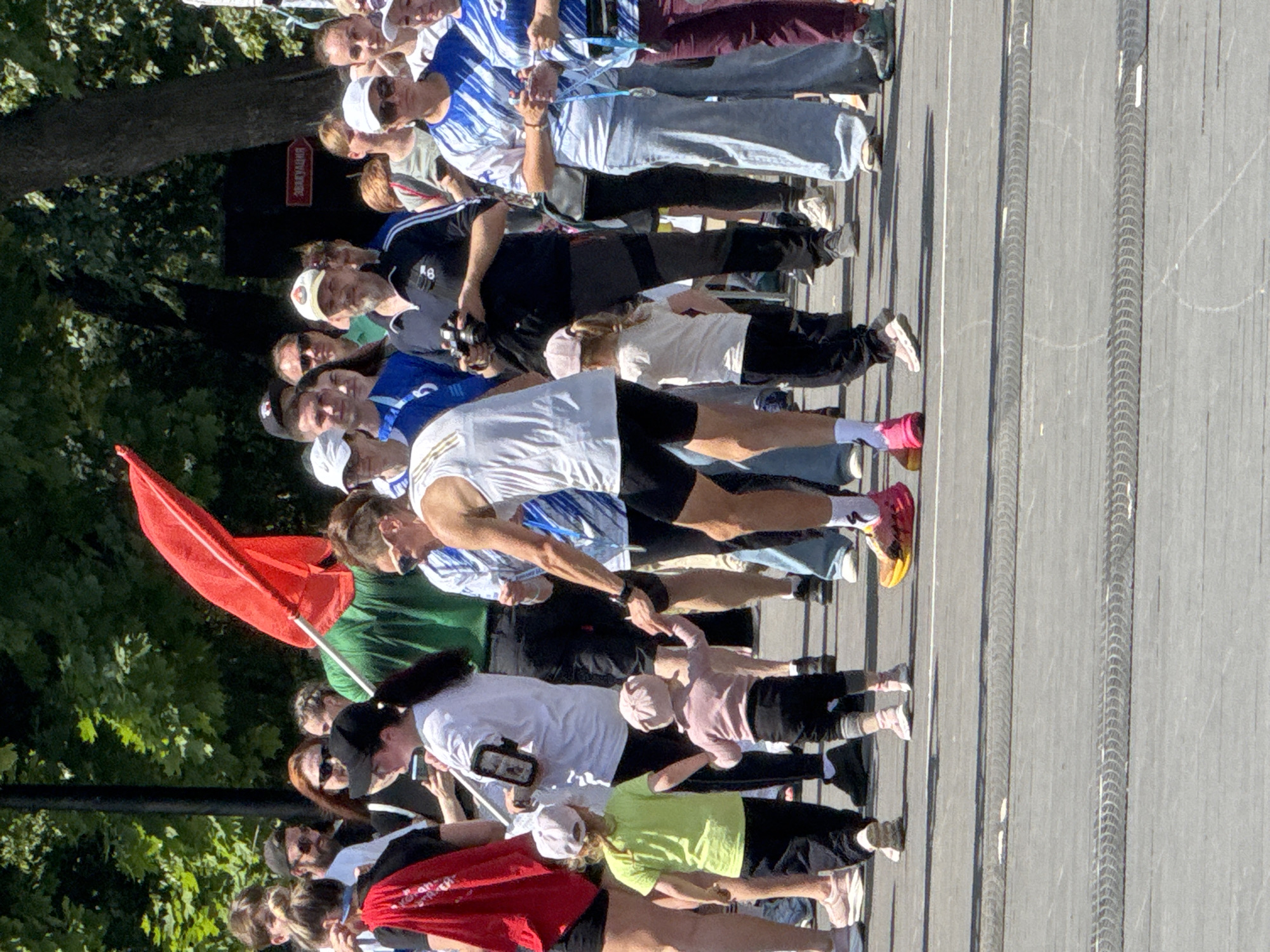 The image shows a crowd of people, including adults and children, on a paved street or plaza. The environment appears to be outdoors with trees in the background and sunlight creating shadows. Some individuals are holding a red flag, suggesting a public gathering or event. A sign in the background, partially visible, shows Russian text, and research indicates it reads "Эвакуация", meaning "Evacuation." This sign, along with common public gathering spaces in Izhevsk, suggests the location is likely a street or public square in Izhevsk, Russia.
Specific individuals visible include a person in the foreground holding a camera, another person in a white tank top and black shorts, and several children. Two young children are in the center of the frame, one in a lime green shirt and black pants, and another in a pink sweater and black pants, appearing to be walking together. A woman in a blue and white striped shirt is also prominent, as are others in casual clothing. The overall scene depicts a dynamic moment within a public gathering.