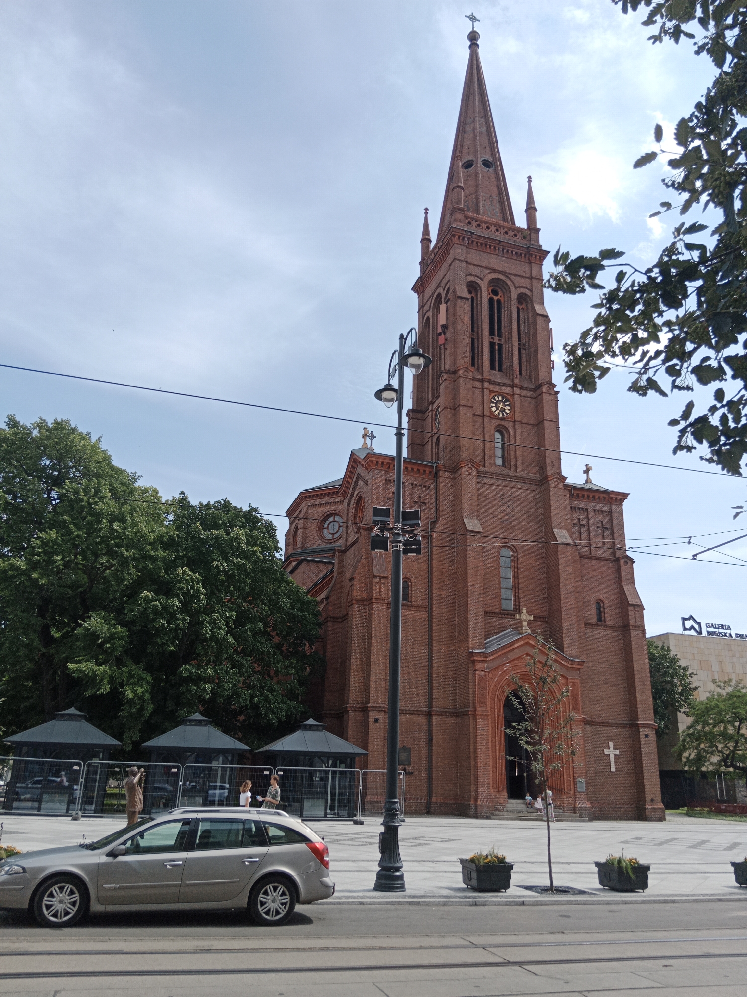 The image shows the brick facade of a church with a tall, pointed steeple. The steeple has Gothic architectural elements and a clock face. The church is surrounded by trees and a paved public square. In the foreground, a silver station wagon is parked on the street. The sky is overcast with some clouds. The church is St. Vincent de Paul Basilica in Bydgoszcz, Poland.